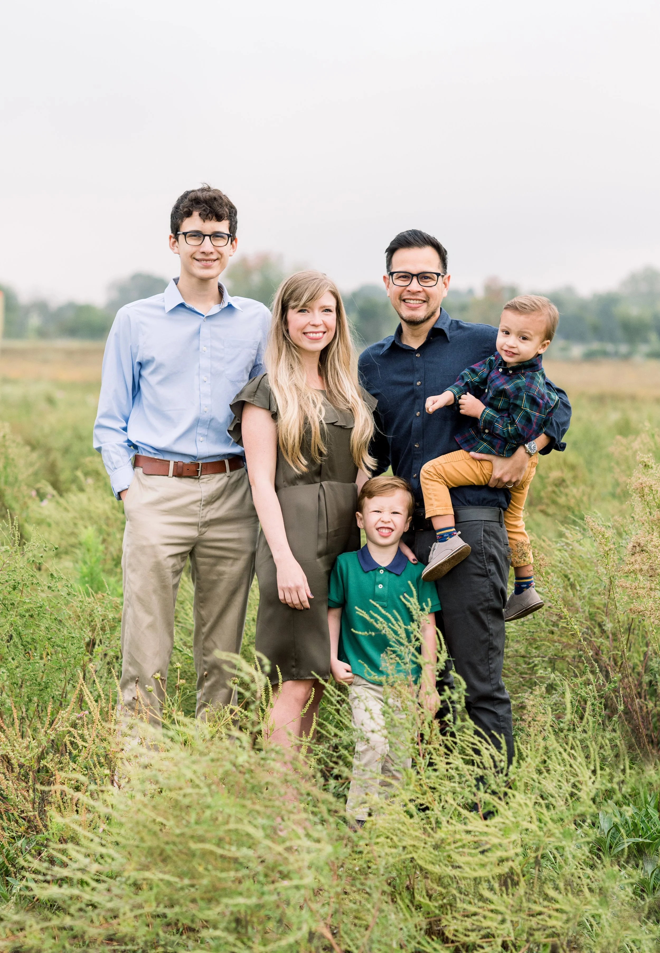 Family of five standing outdoors in a field during daytime, smiling at the camera.