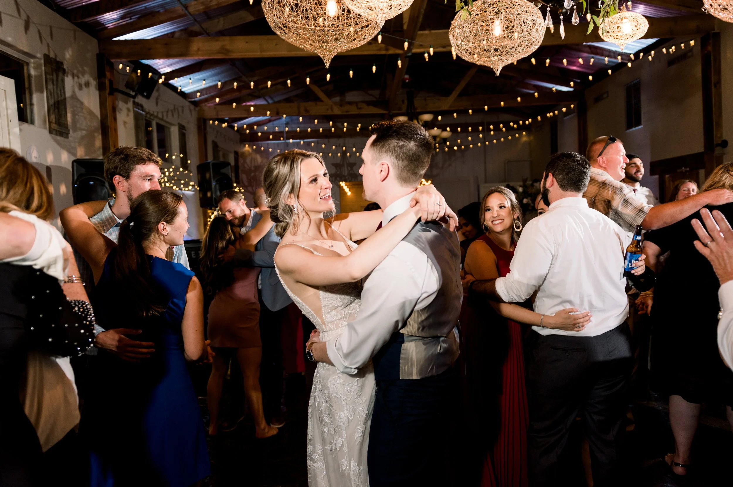 Couple dancing at a wedding reception with guests around them, string lights and elegant chandeliers overhead.