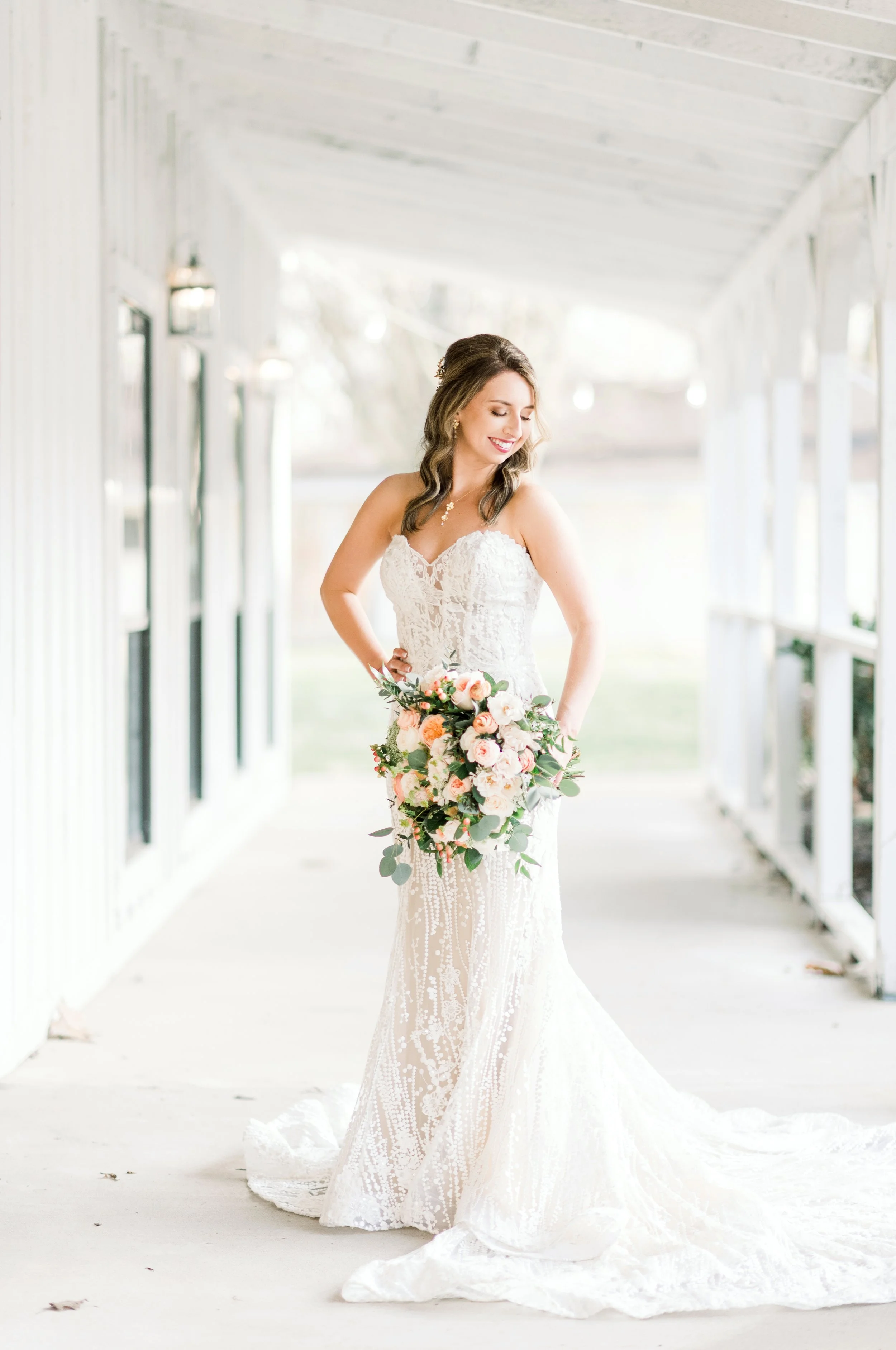 A bride in a white lace wedding dress holding a bouquet of pink and white flowers, standing on a porch with a white ceiling and walls, smiling and looking down.