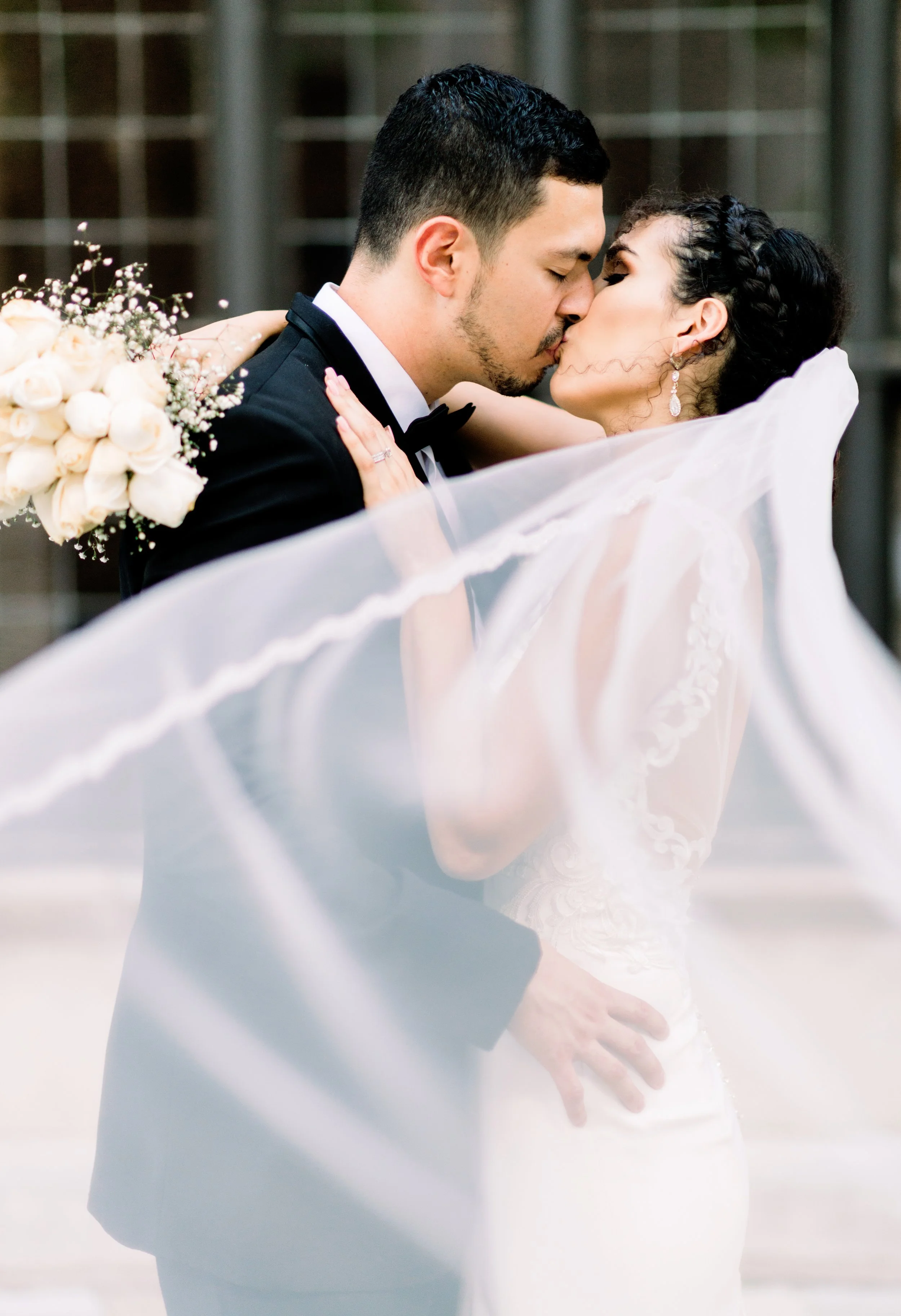 A bride and groom sharing a kiss, with the bride's veil flowing in the foreground, holding a bouquet of white roses with baby's breath, outdoors near a brick building.