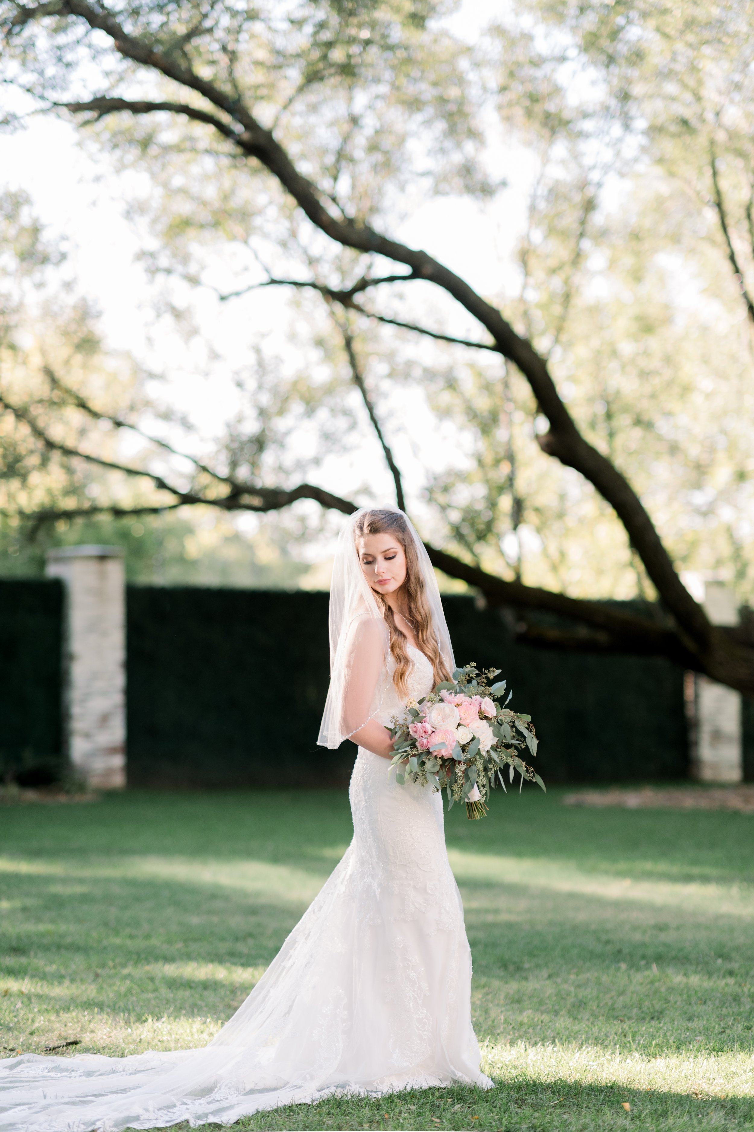 Bride in a white lace wedding dress holding a bouquet of pink and white flowers outdoors in a garden with green grass and trees.