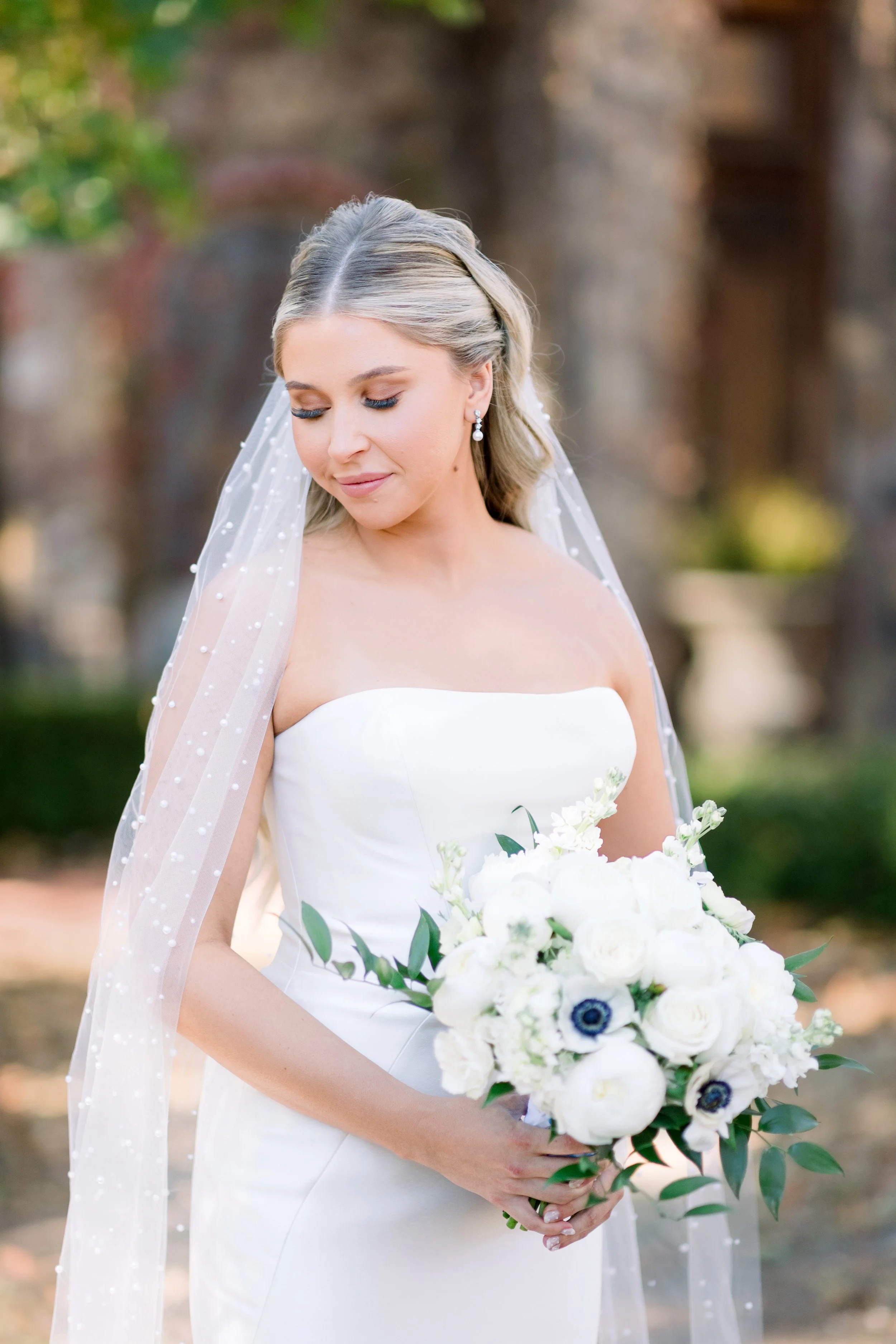 Bride wearing a white strapless wedding dress holding a bouquet of white flowers and greenery, outdoors with blurred trees and background.