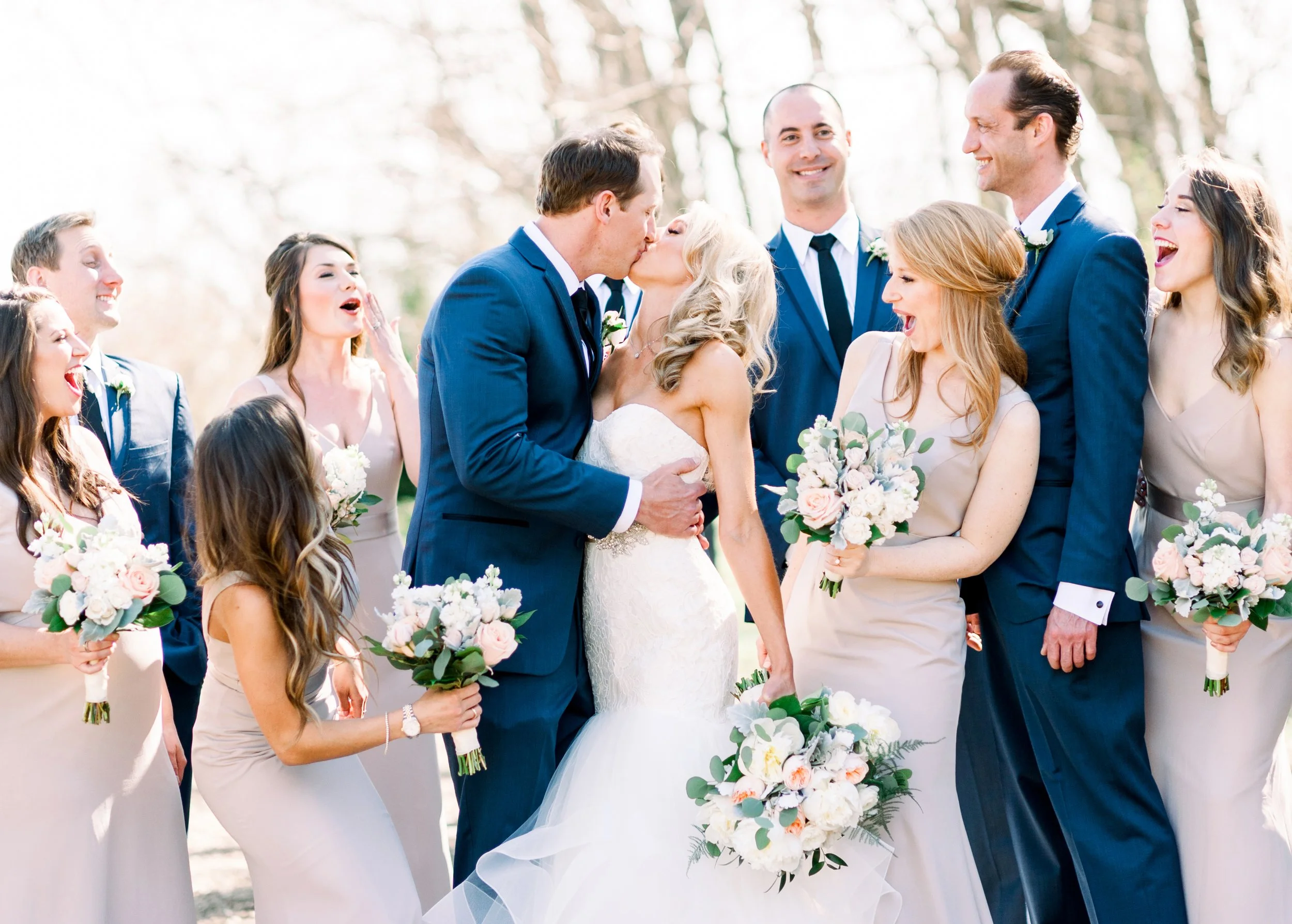Wedding celebration featuring a couple kissing, surrounded by bridesmaids and groomsmen outdoors on a sunny day.