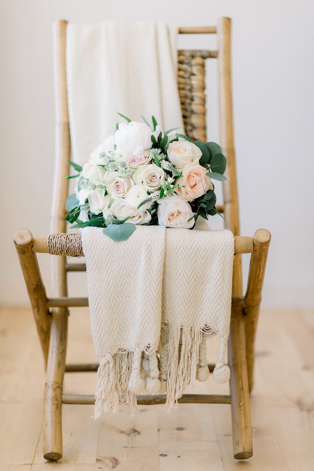 A bouquet of white and blush roses with greenery resting on a wooden chair with a white textured scarf draped over it.