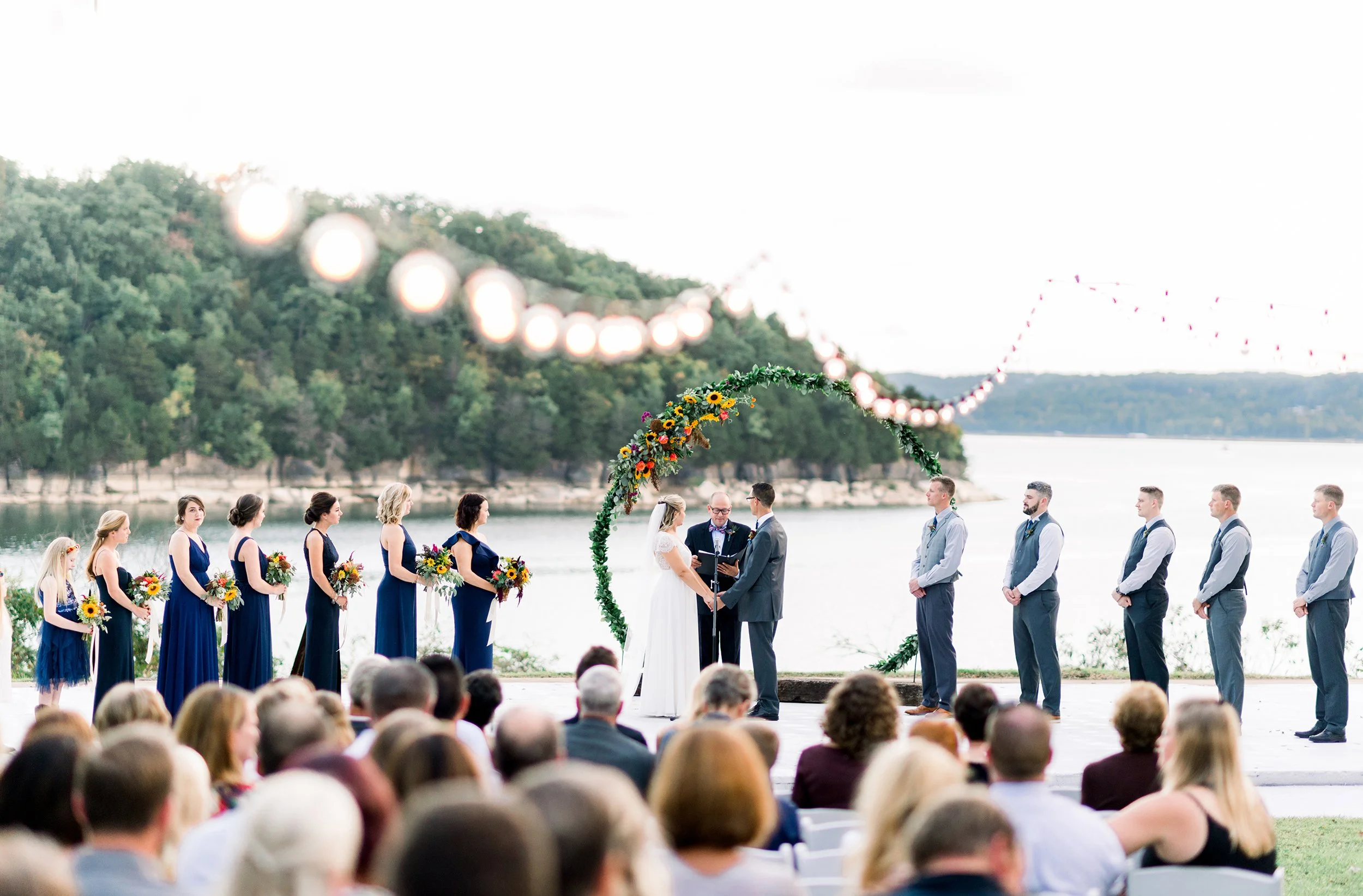 Outdoor wedding ceremony by a river with the bride and groom exchanging vows under a green floral arch, with bridesmaids and groomsmen standing beside them, and an audience seated in front, during daytime with trees and hills in the background.