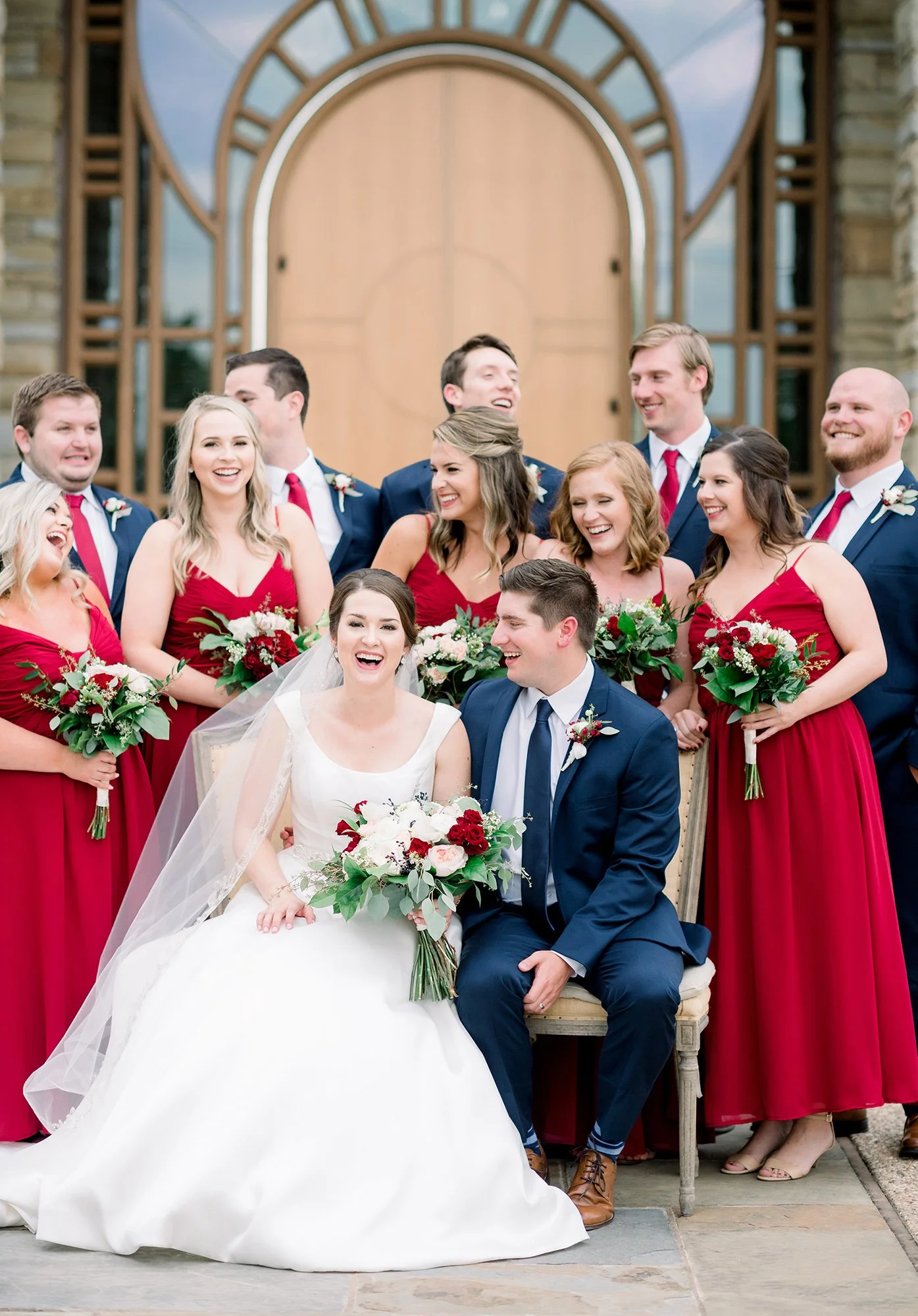 A wedding party posing outdoors, featuring a bride in a white gown and veil, holding a bouquet of white and red flowers, and a groom in a navy suit with a boutonnière, surrounded by bridesmaids in red dresses and groomsmen in navy suits with red ties