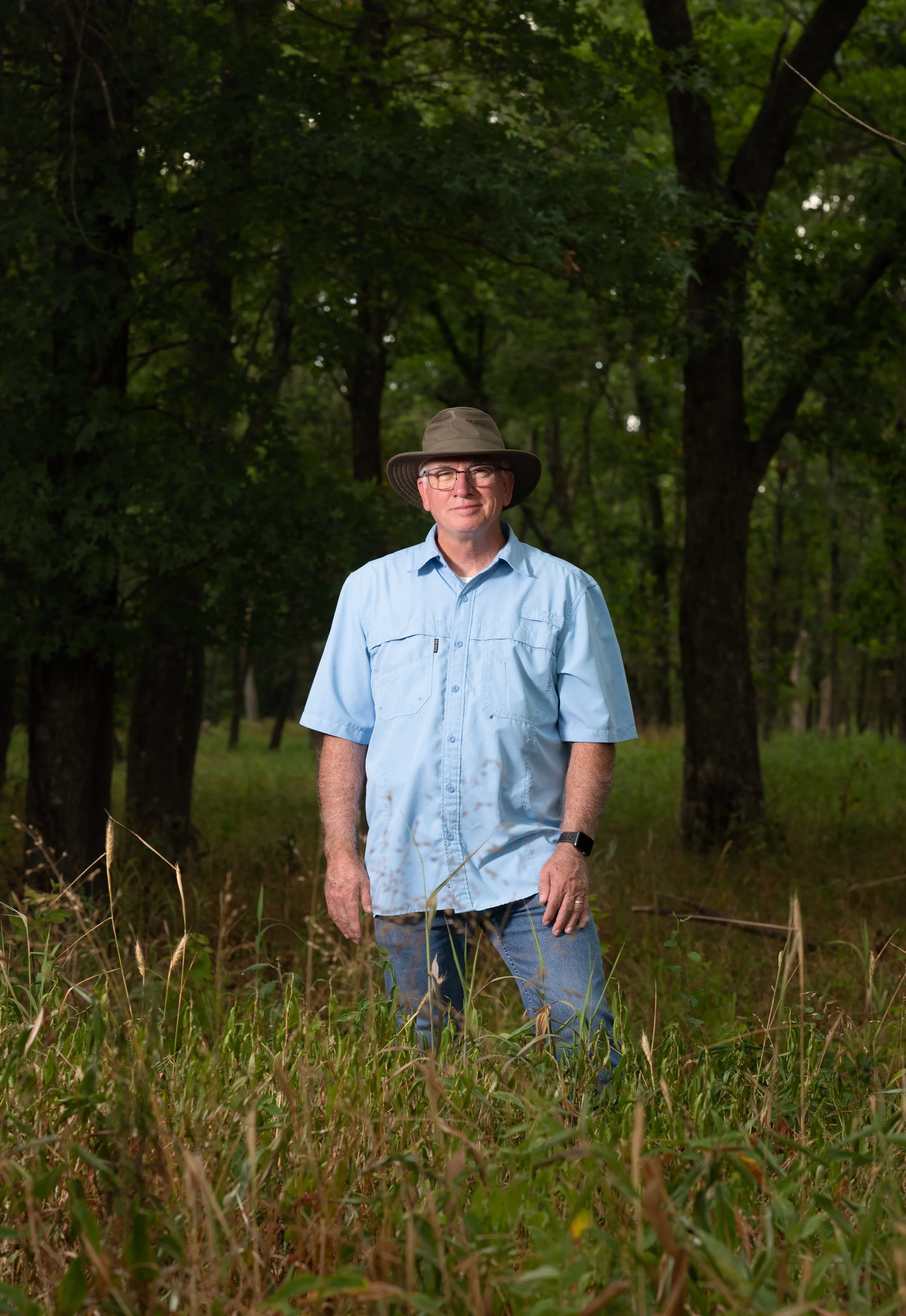 A man wearing a wide-brimmed hat, glasses, a light blue short-sleeved shirt, and jeans, standing in a grassy, wooded area.