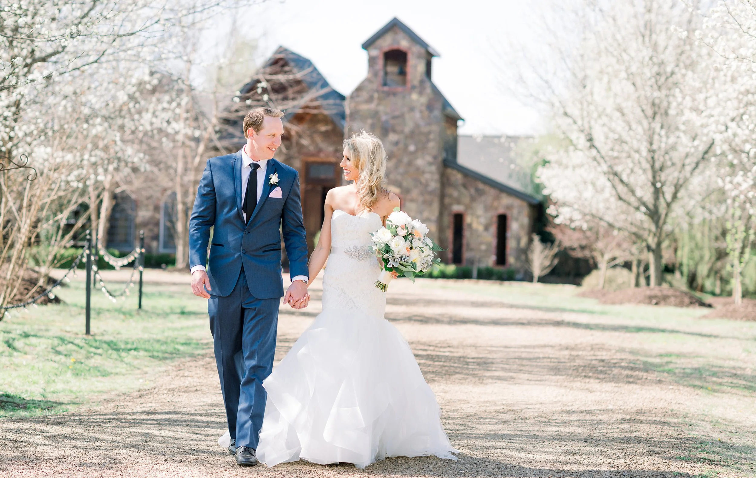 A bride and groom walking hand in hand outside on a sunny day, with blooming trees and a brick church in the background, celebrating their wedding.