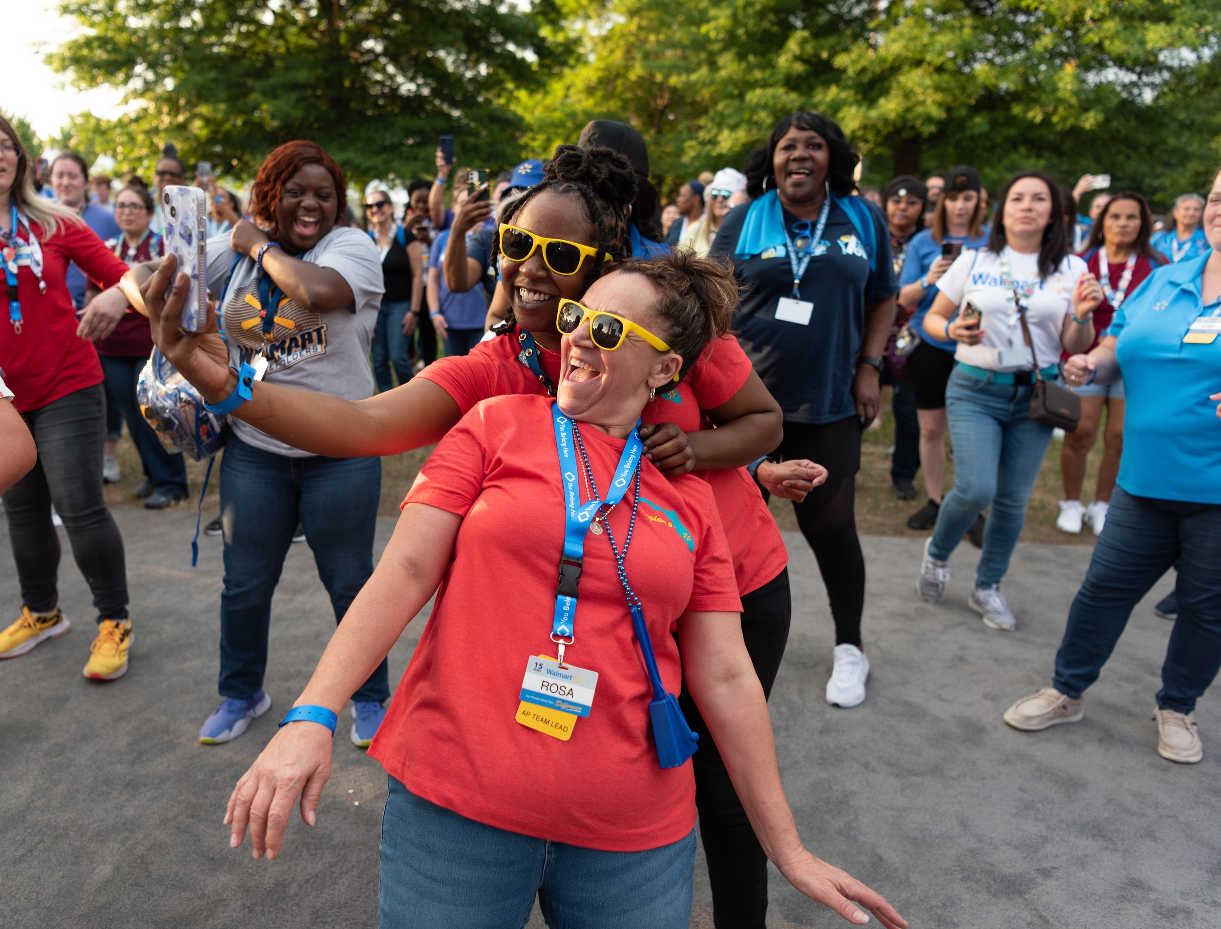 Group of diverse women dancing and taking selfies outdoors during a joyful event, with trees in the background.