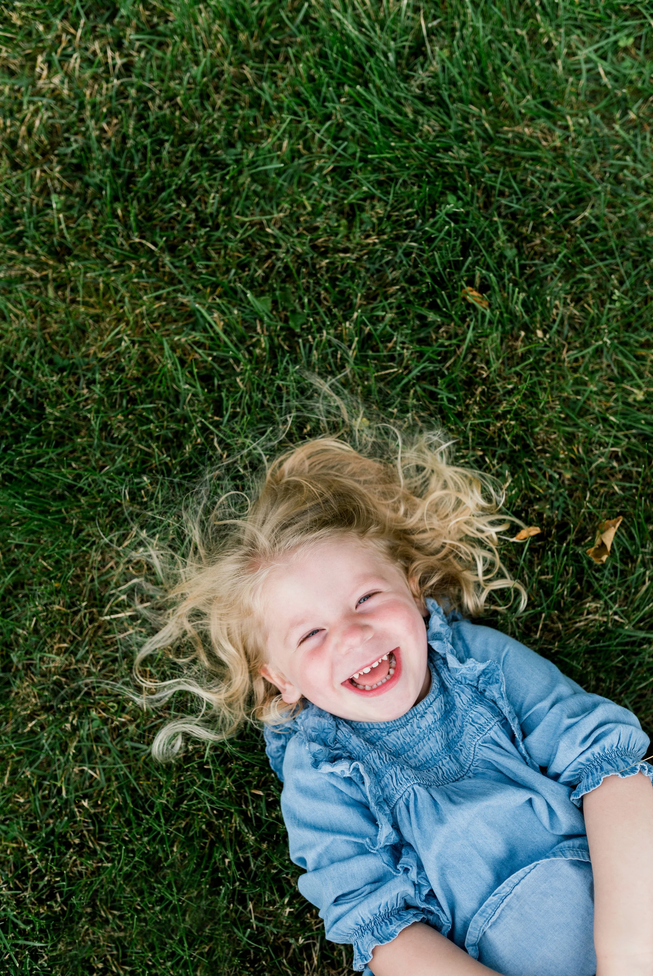 A young girl with curly blonde hair lying on green grass, smiling and laughing.