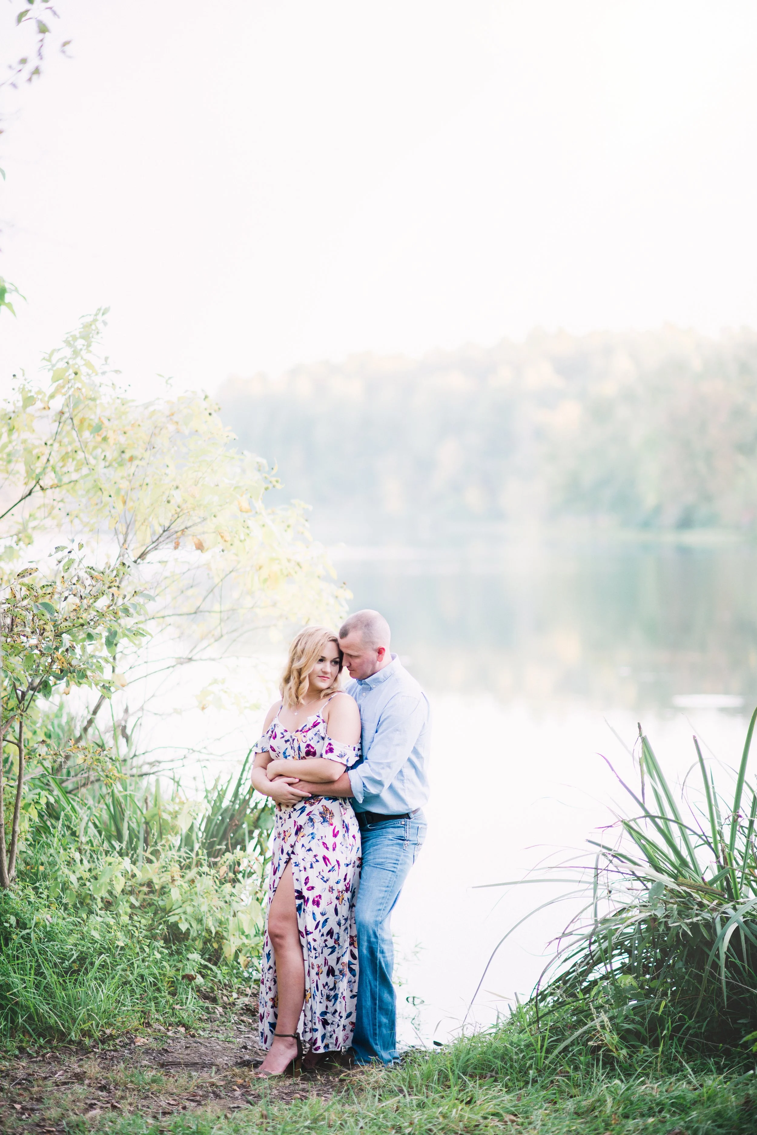A couple standing by a lake, embracing, with trees and water in the background on a bright day.