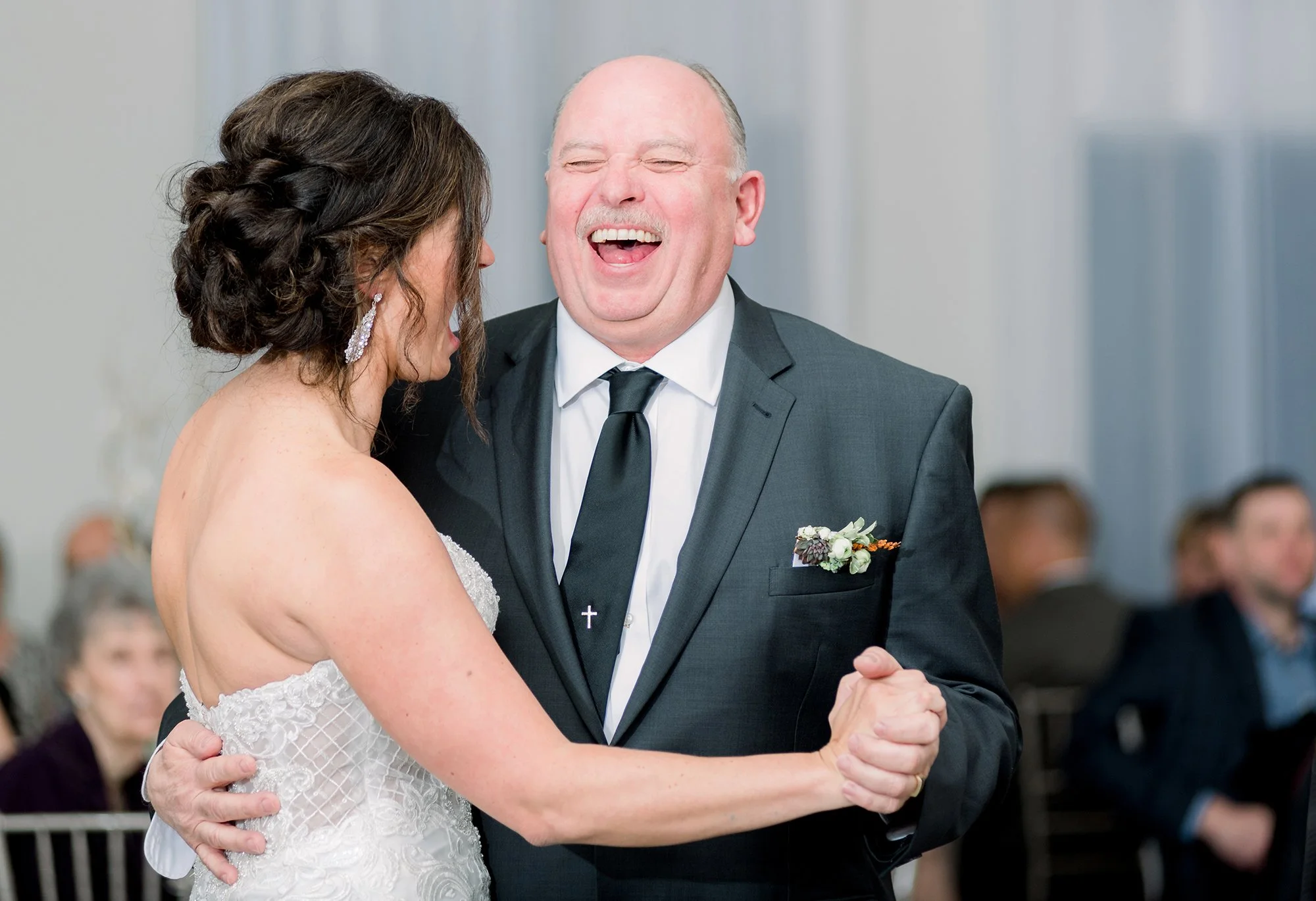 A bride and an older man, possibly her father, dancing and smiling at a wedding reception. The bride is wearing a white lace wedding gown and the man is in a dark suit with a boutonniere.