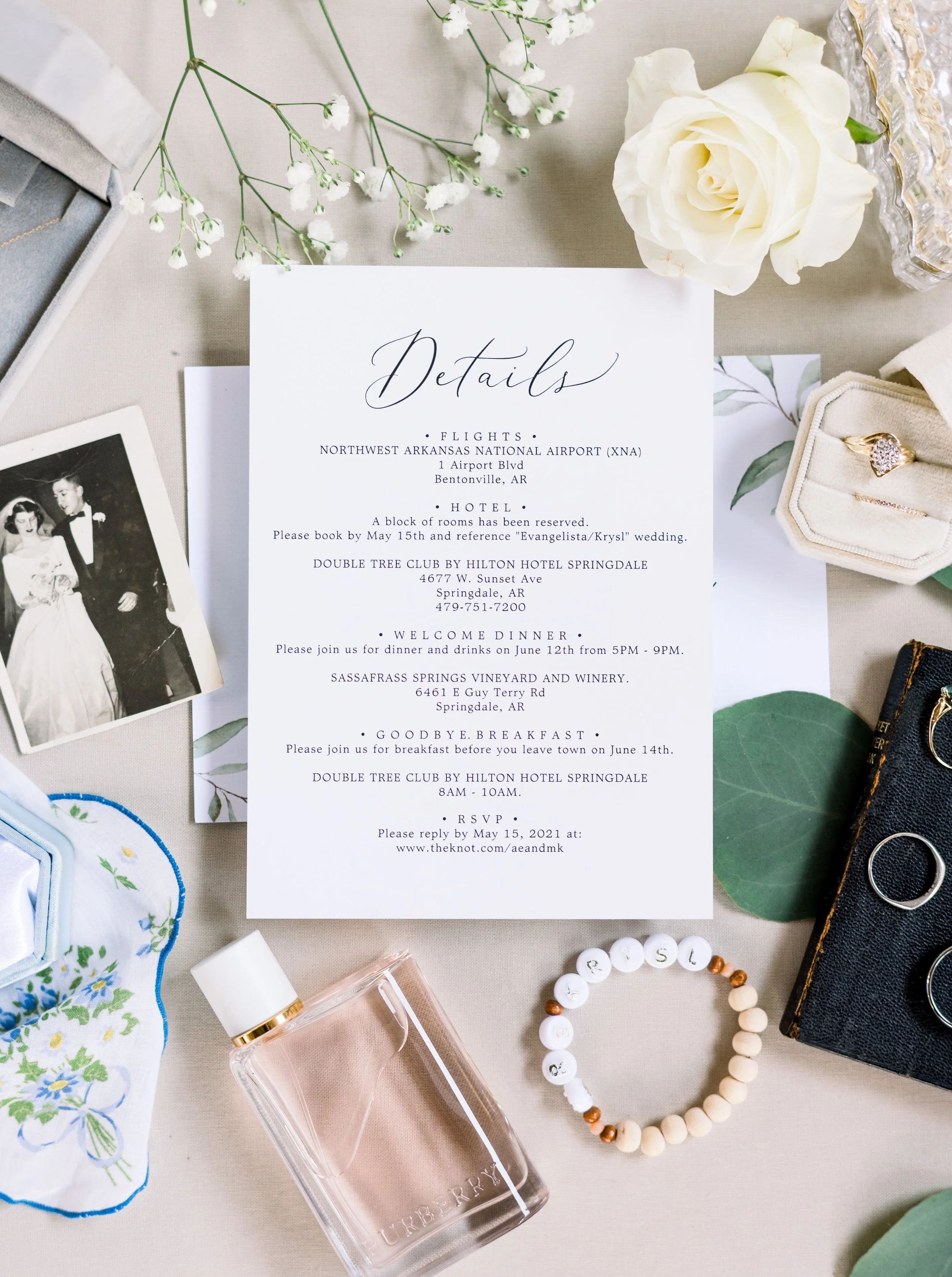 Wedding details invitation surrounded by flowers, jewelry, a vintage photograph, perfume, and other accessories.
