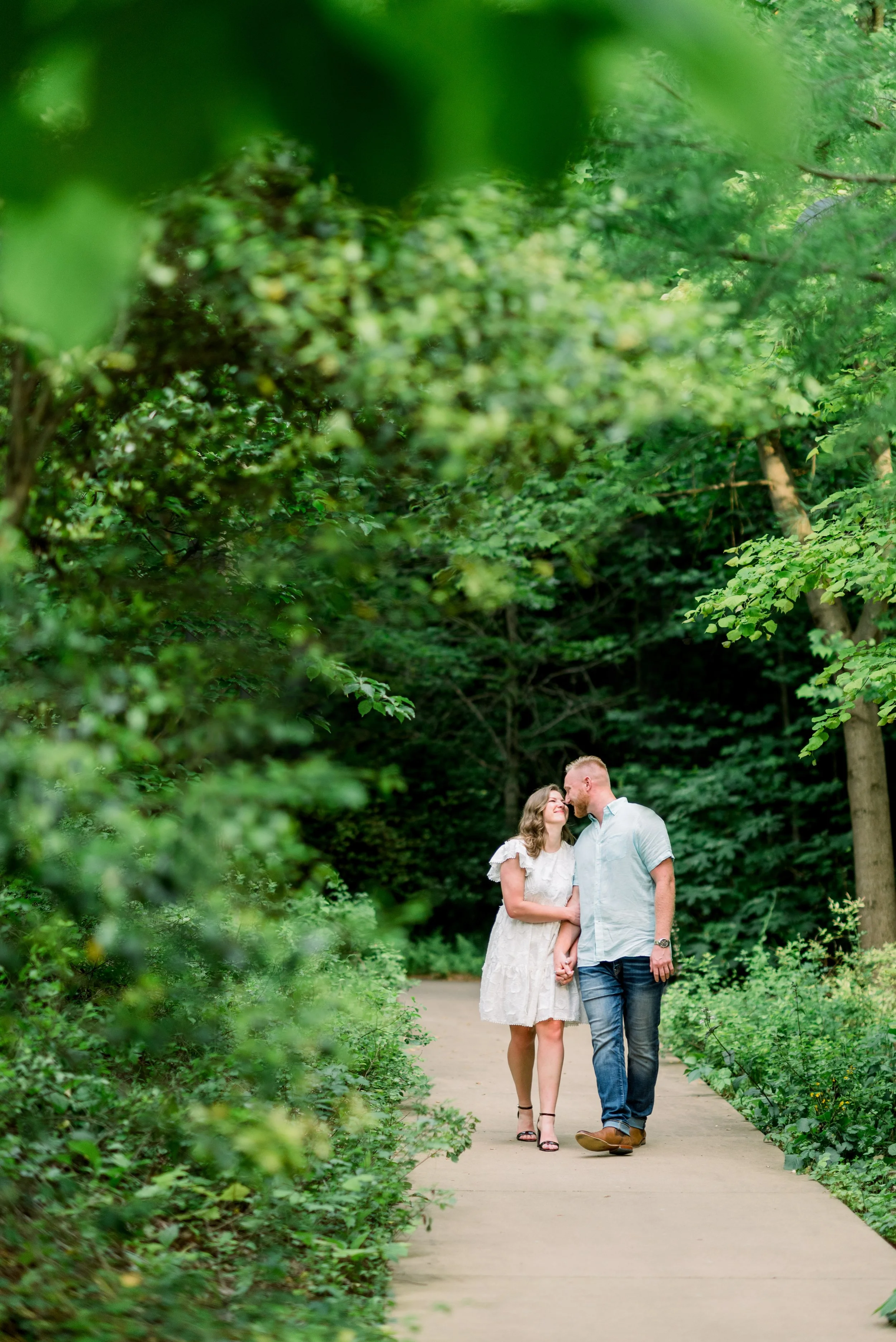 A couple holding hands and sharing a kiss while walking on a path through a lush green forest.