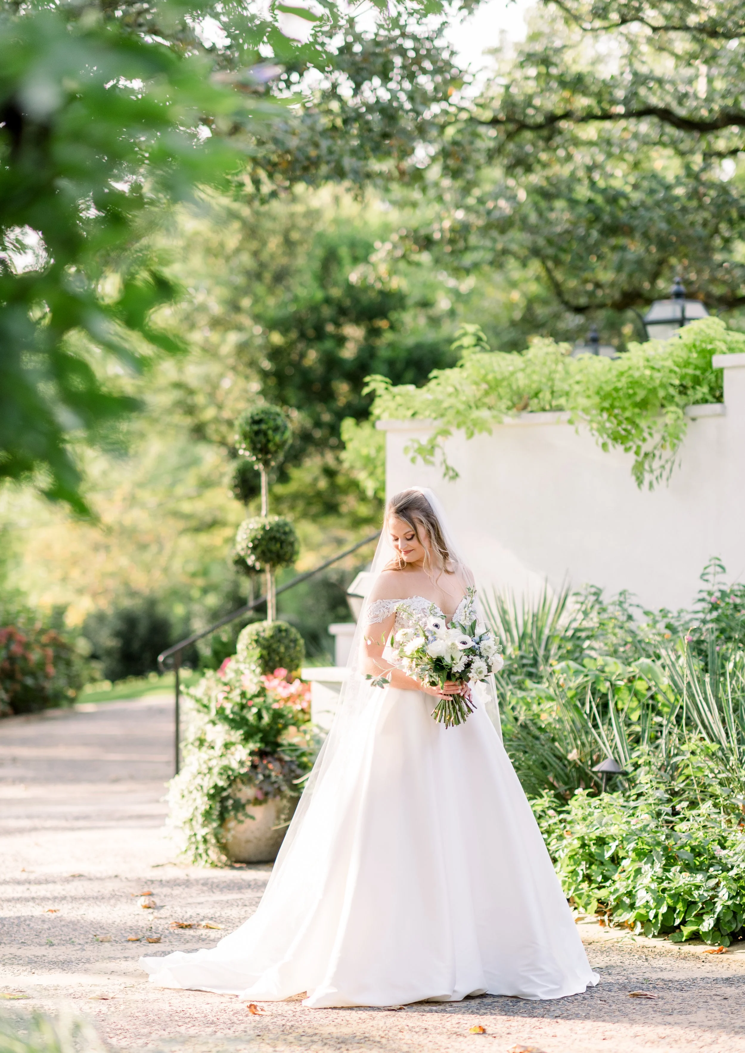 A bride in a white wedding dress holding a bouquet of white flowers, standing outdoors surrounded by greenery and decorative plants, with sunlight filtering through trees.