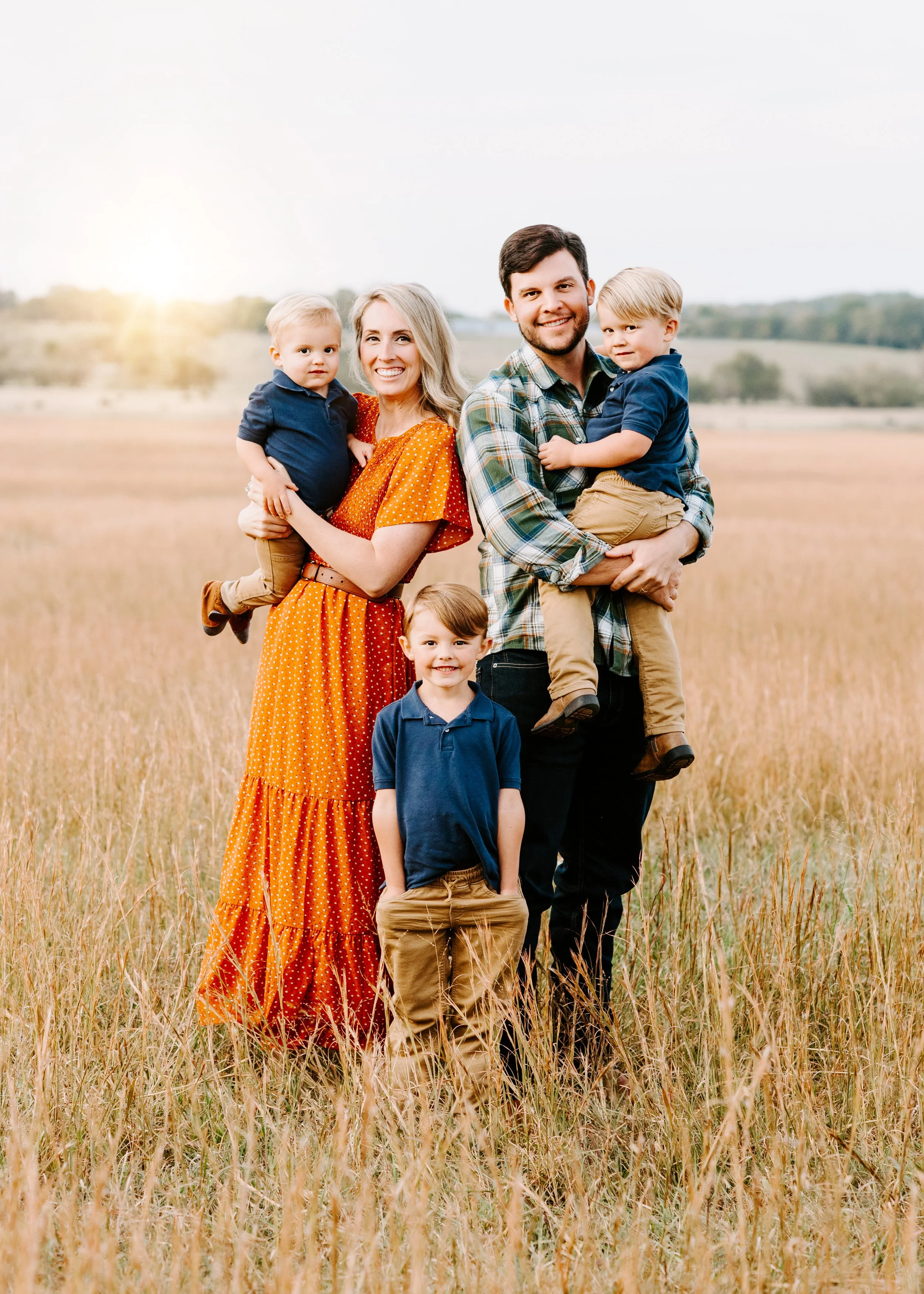A family of five standing in a field during sunset, with two young boys and a woman holding two more children, all smiling at the camera.