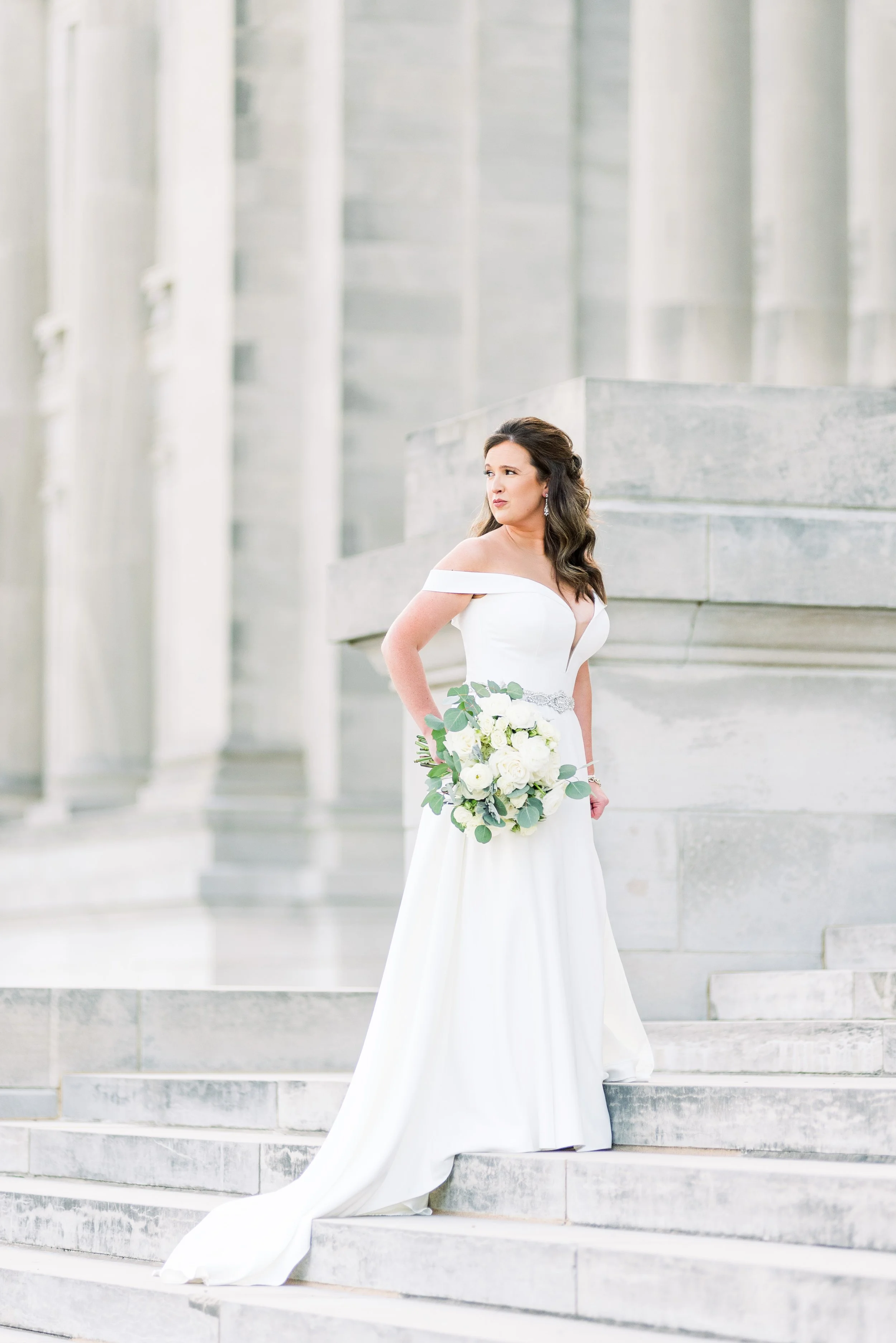 Bride in an off-shoulder white wedding gown holding a bouquet of white roses and greenery, standing on stone steps outside a building with tall columns.