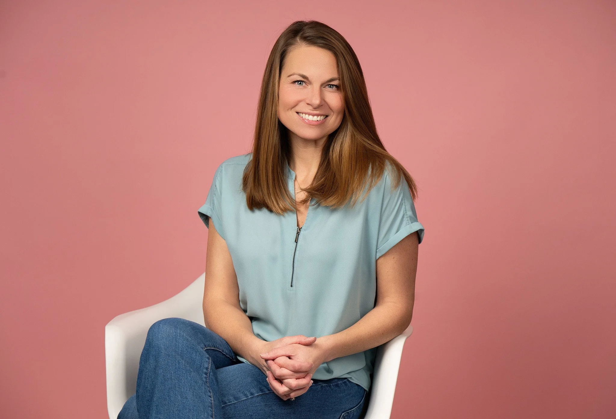 A woman with long brown hair, wearing a light blue top and jeans, sitting on a white chair against a pink background, smiling at the camera.