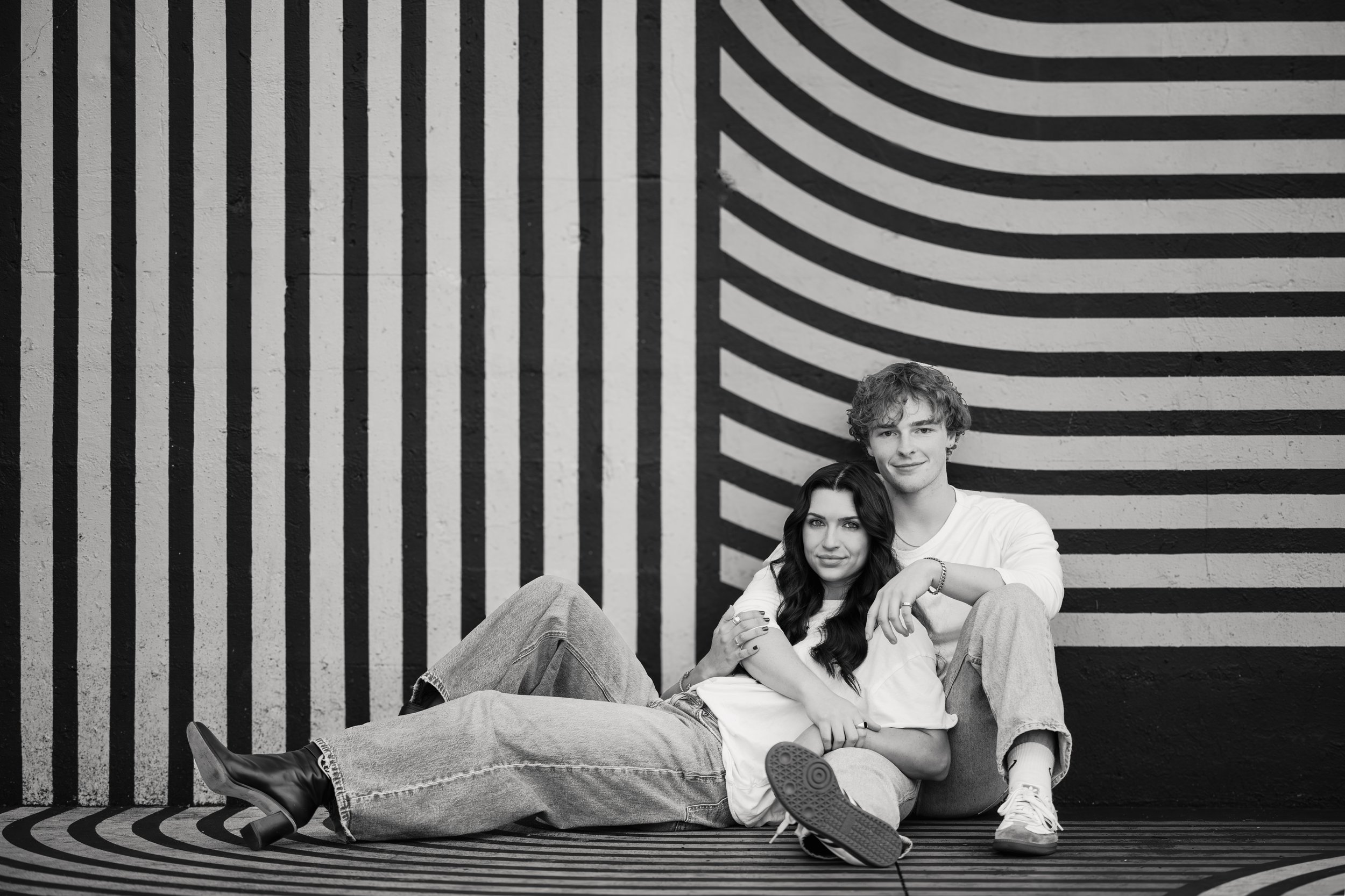 A young man and woman sitting on the ground in front of a wall with black and white geometric stripes and patterns. The woman is lying down, leaning on the man's lap, and the man is sitting with one knee up.