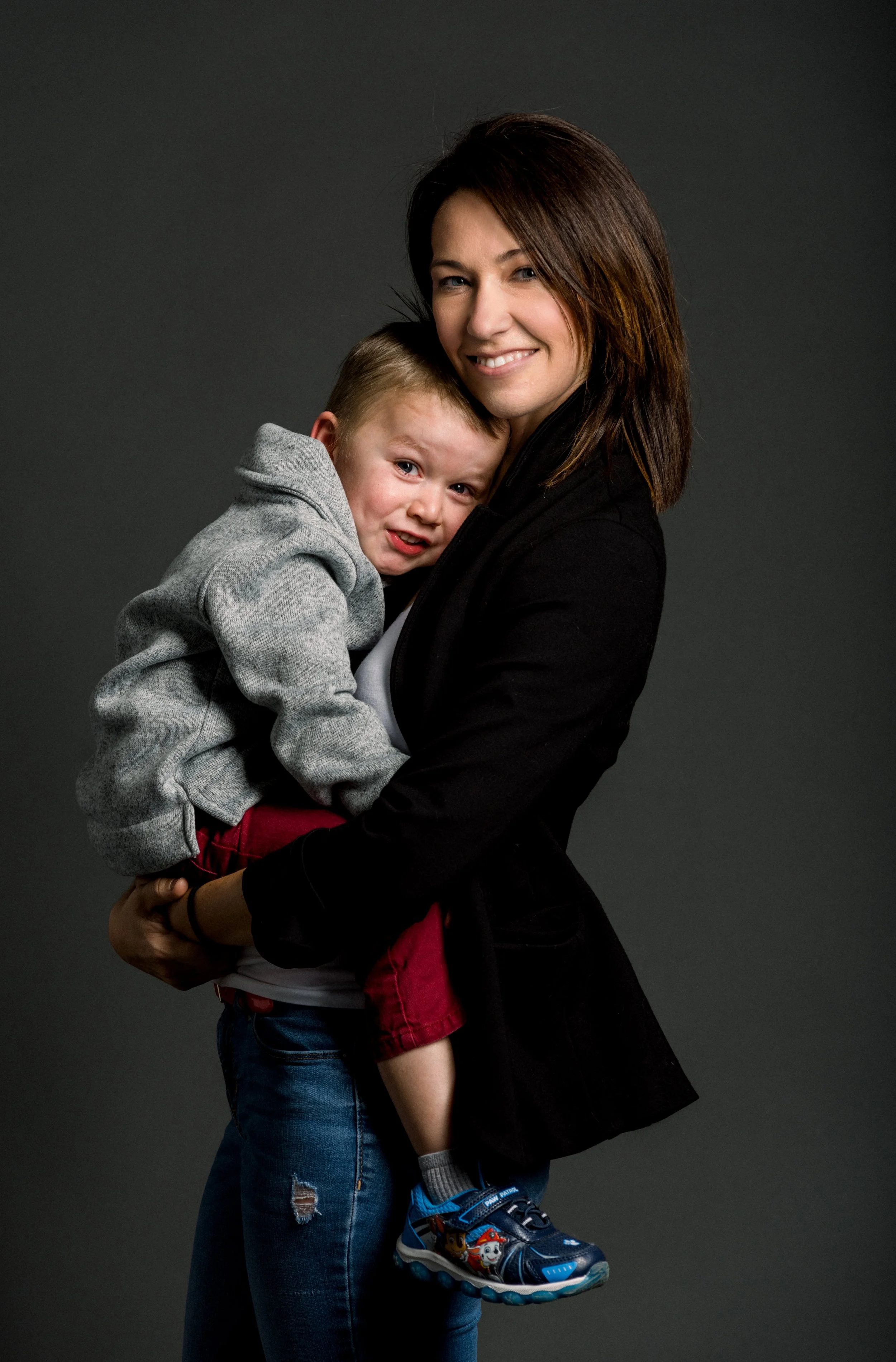 A woman holding a young boy in her arms, both smiling, against a dark background.