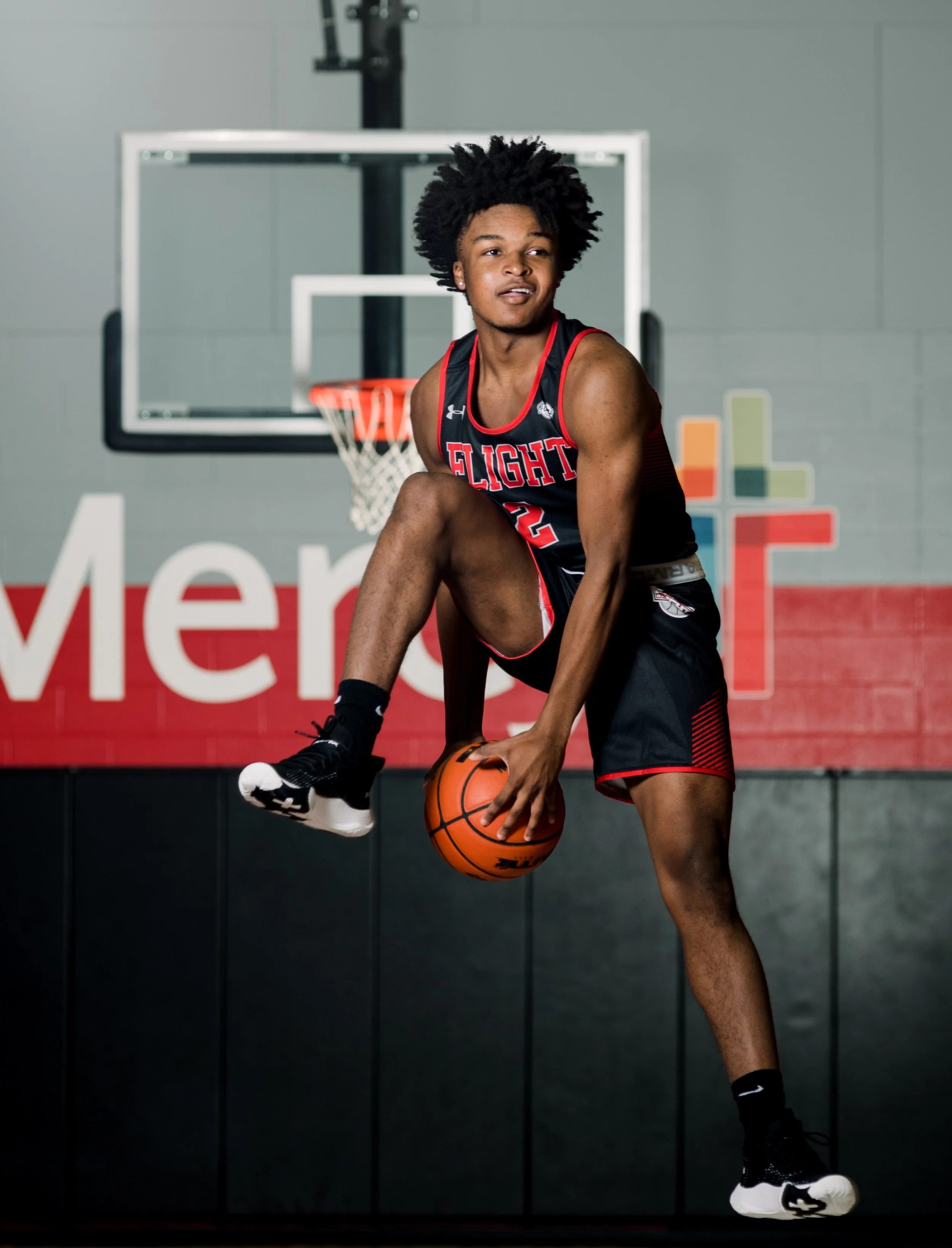 Young male basketball player in uniform practicing on indoor court with basketball hoop in background.