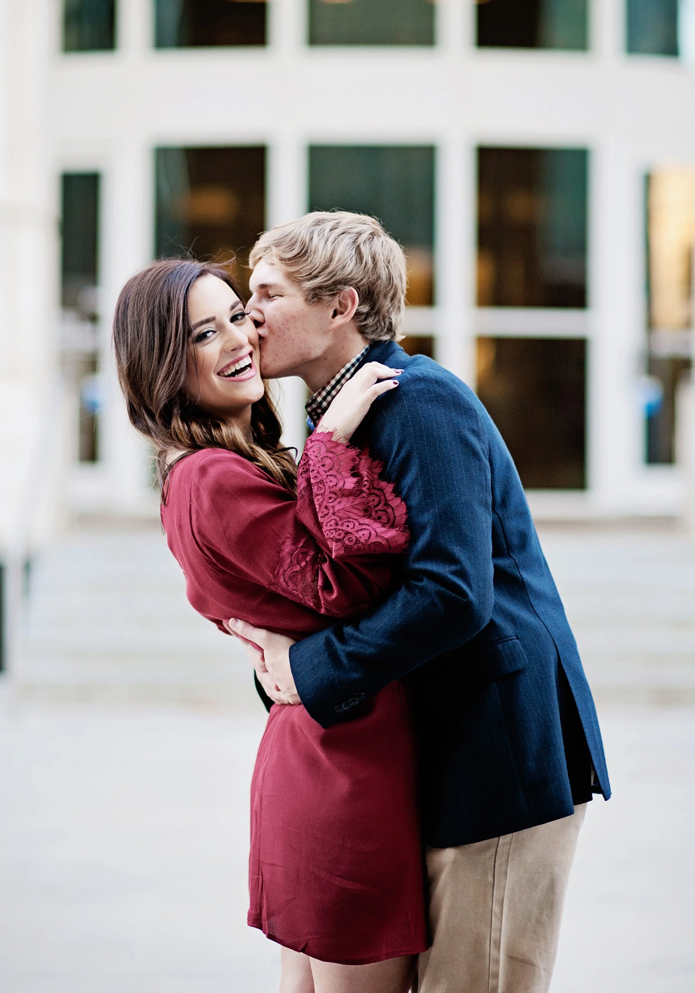 A couple sharing a kiss, with the man in a dark blue blazer and khaki pants and the woman in a red dress with lace sleeves, standing outdoors in front of a building with large windows.