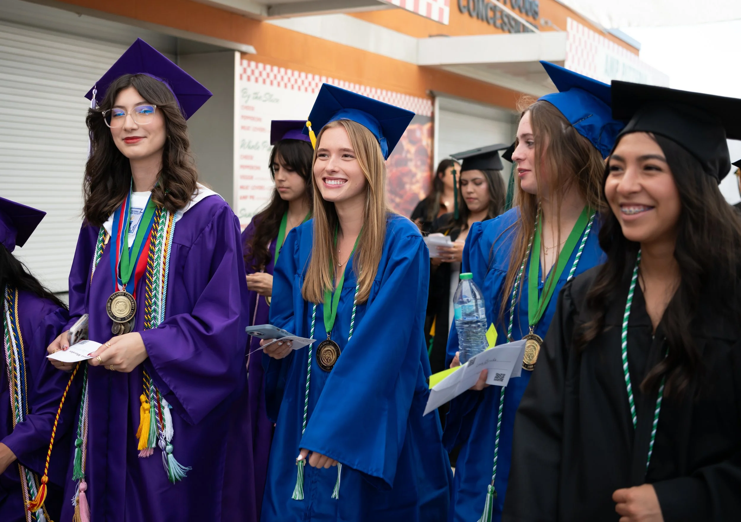 Group of diverse female graduates in purple, blue, and black caps and gowns, wearing honor cords and medals, standing outdoors during graduation ceremony, some smiling and holding diplomas, in front of a building with a banner.