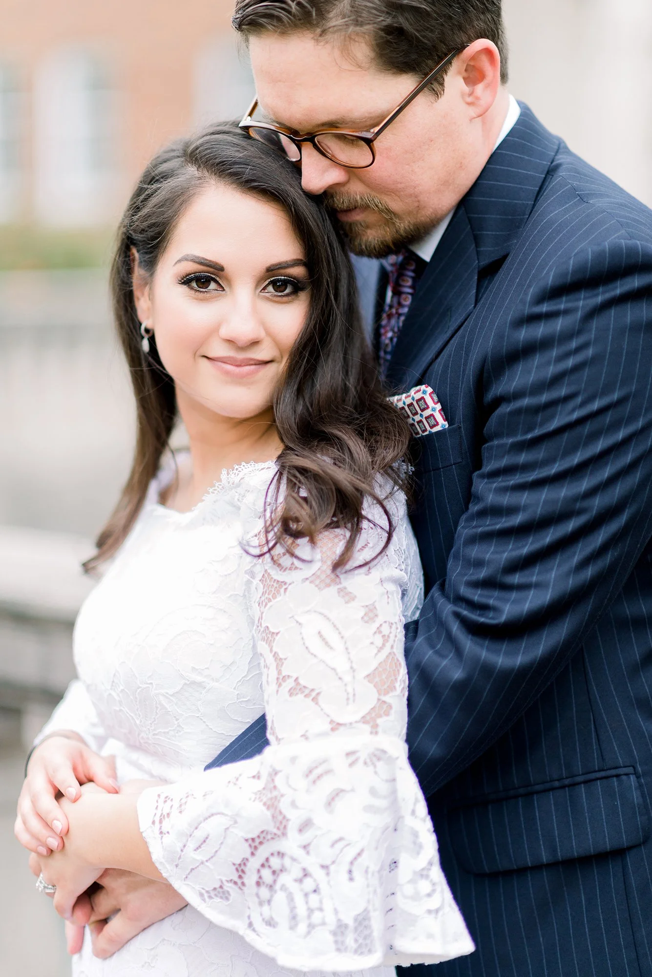 A woman in a white lace dress and a man in a navy pinstripe suit embrace outdoors, with the man gently resting his forehead on her head.