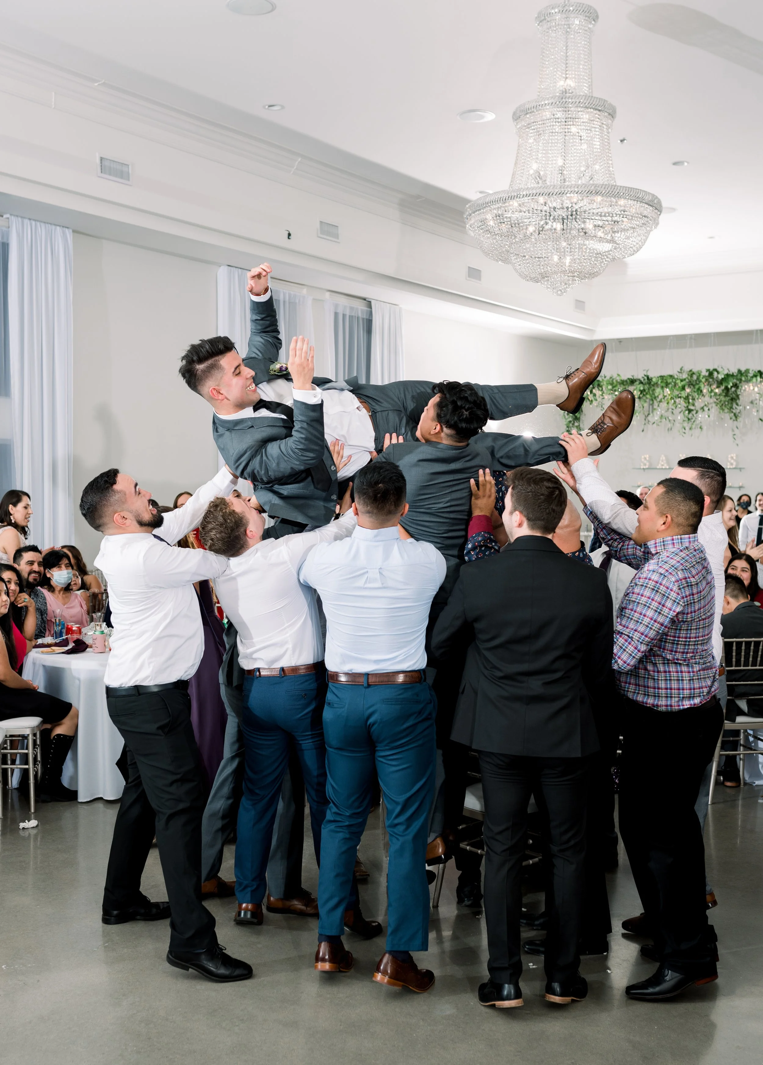 Group of people celebrating at a wedding reception, throwing a man into the air, with onlookers at tables in the background.