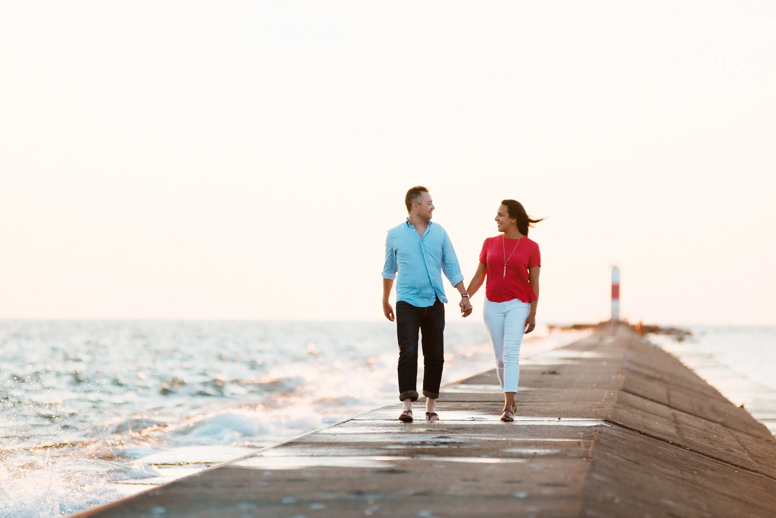 A couple holding hands while walking on a pier next to the ocean with a lighthouse in the background during sunset.