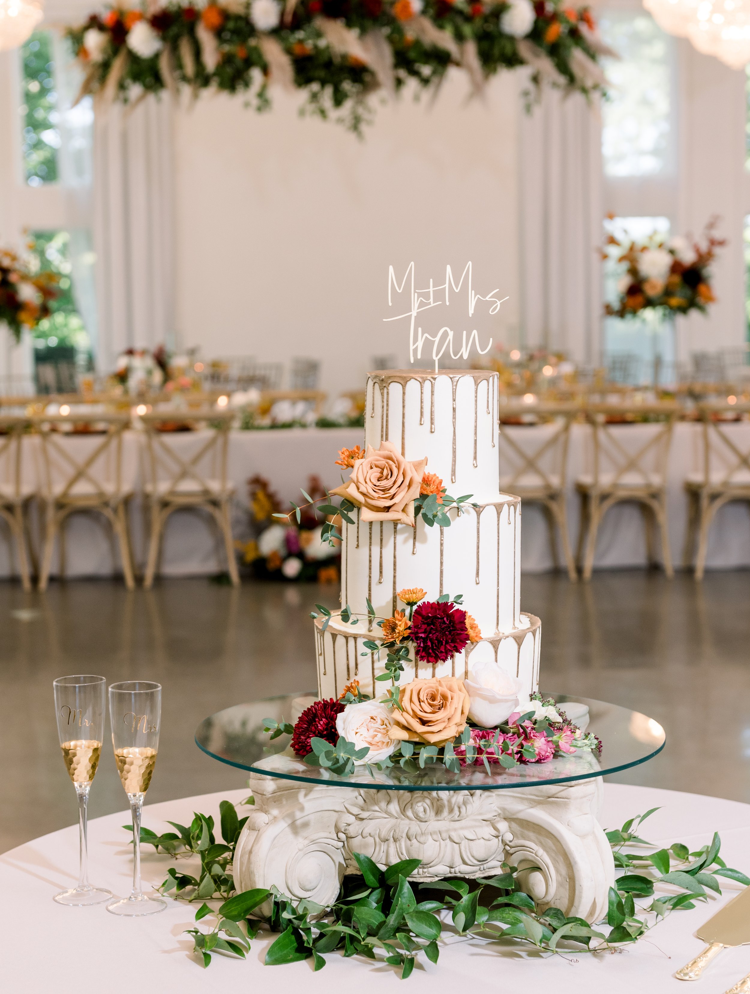 Three-tiered wedding cake with gold drip decoration, adorned with peach and burgundy flowers, eucalyptus leaves, and a topper that reads 'Mr & Mrs Tran'; cake is on a decorative stand with a round mirror surface, surrounded by green leaves, set on a 