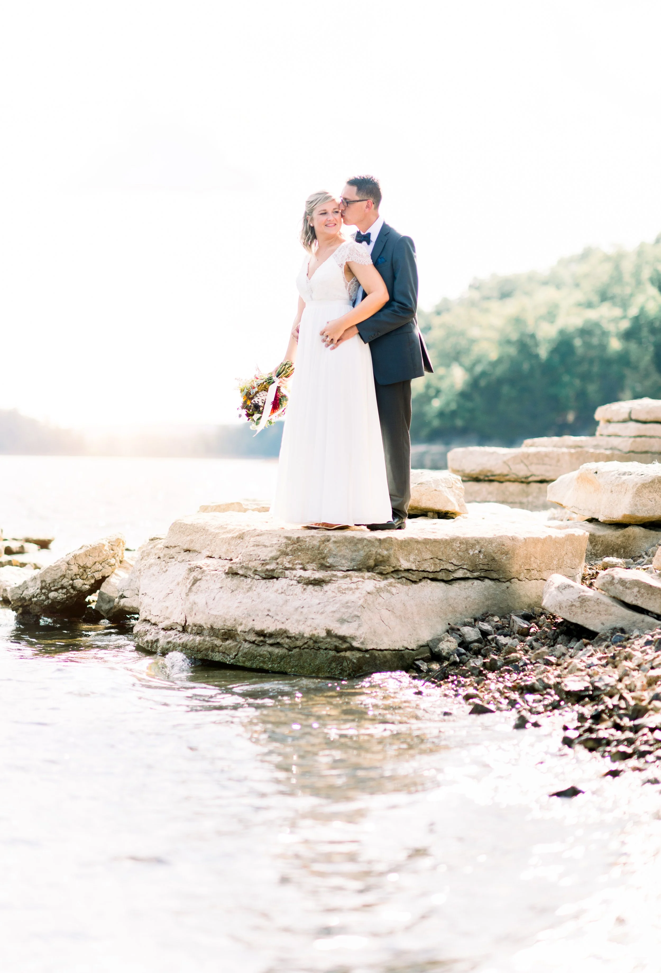 A newlywed couple stands on a large rock by the water, with the groom kissing the bride on the cheek and the bride holding a bouquet, outdoors during daylight.