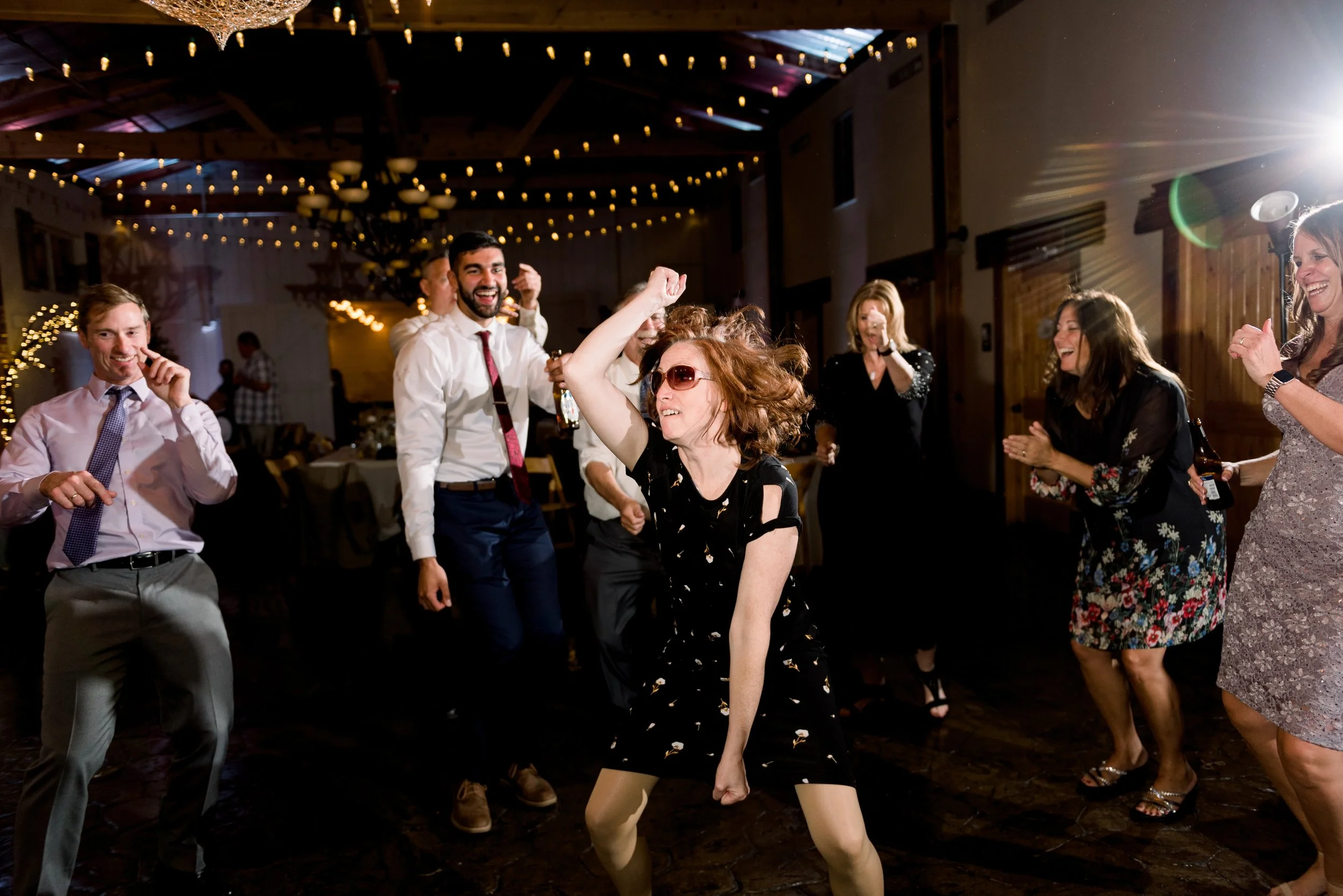 People dancing and celebrating at a wedding reception or party in a decorated venue with string lights and wooden walls.