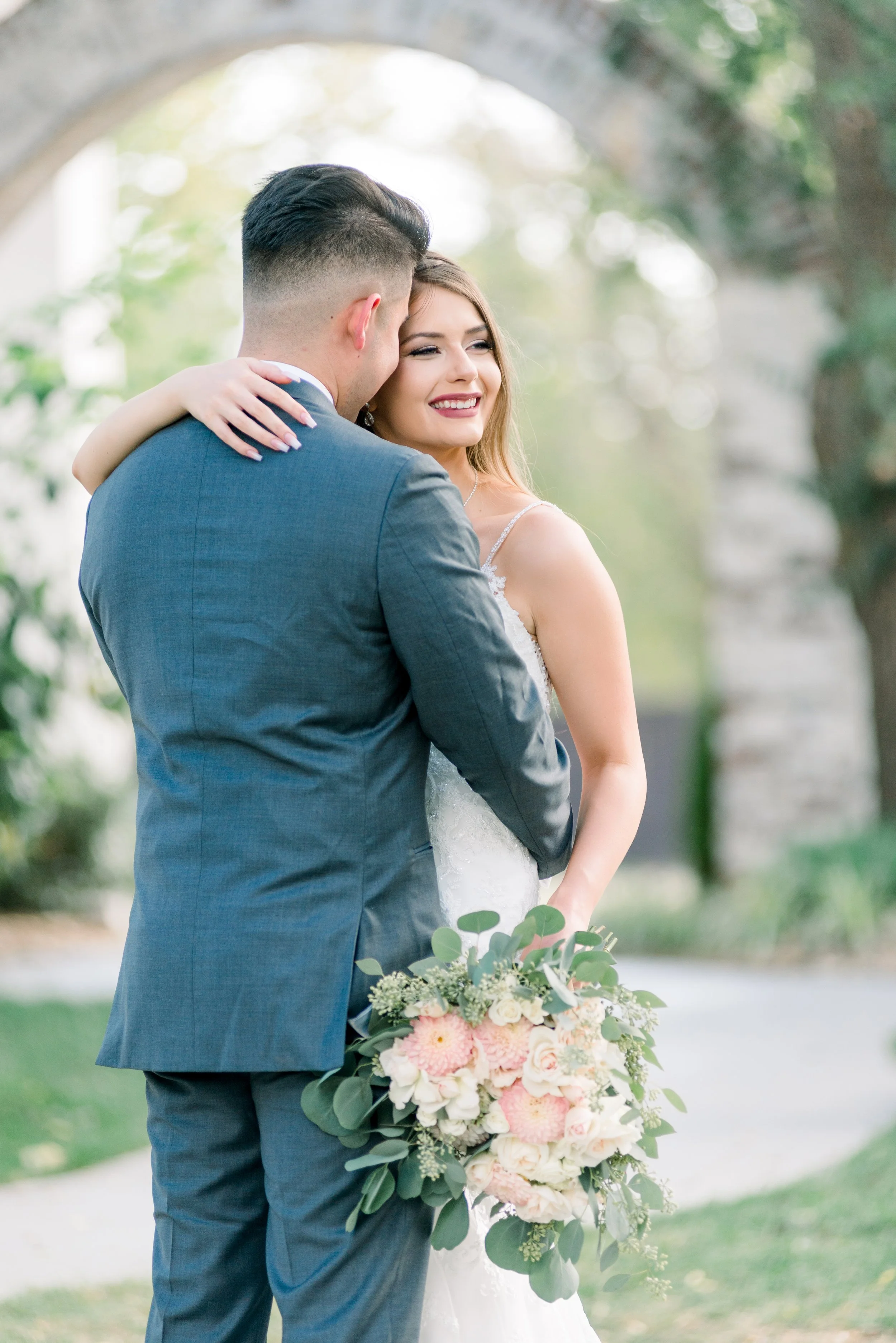 A bride and groom embrace outdoors, with the bride holding a bouquet of pink and white flowers, showing happiness.