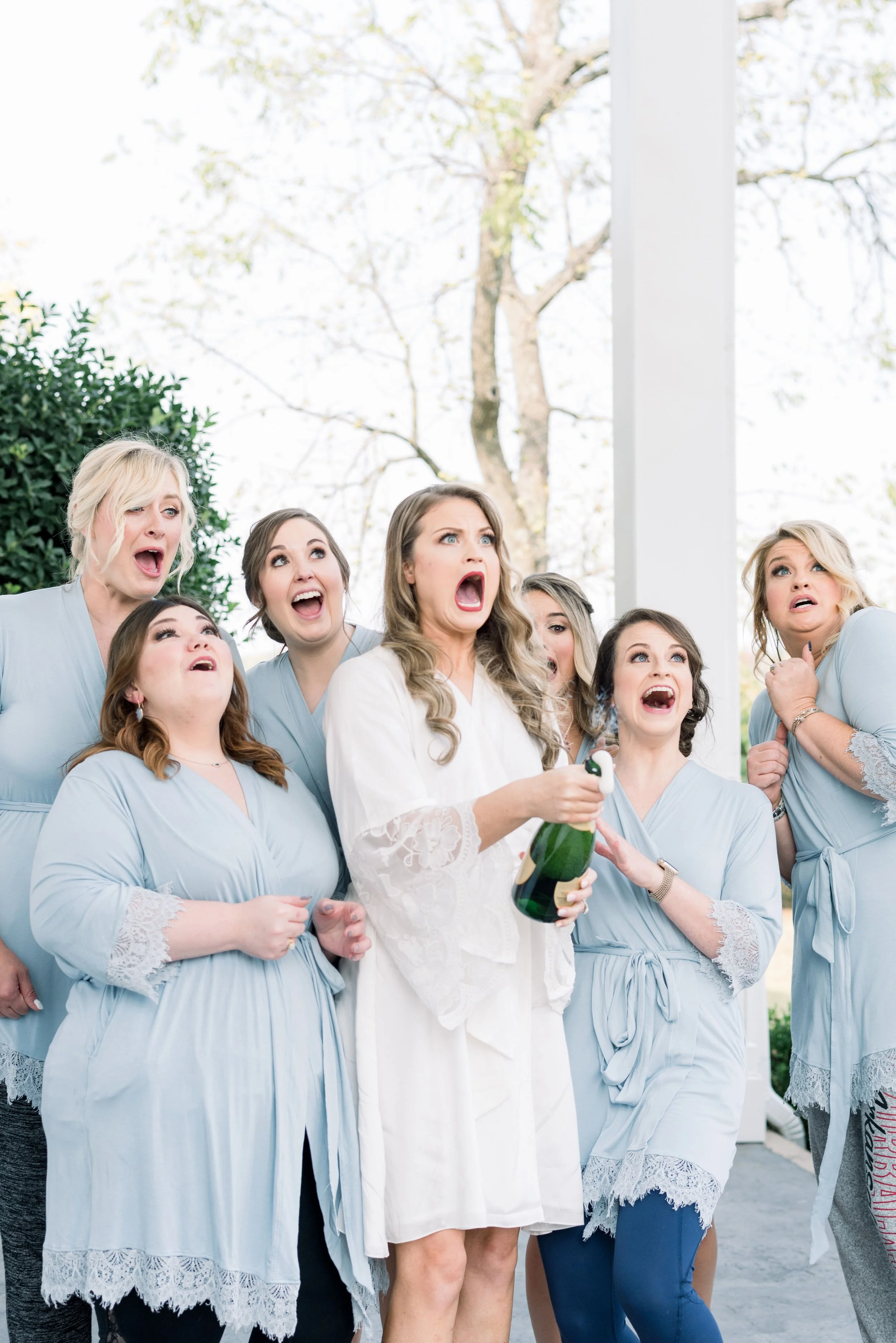 A group of women in light blue gowns attending a wedding, showing surprise or shock, with one woman in a white robe holding a bottle of champagne.