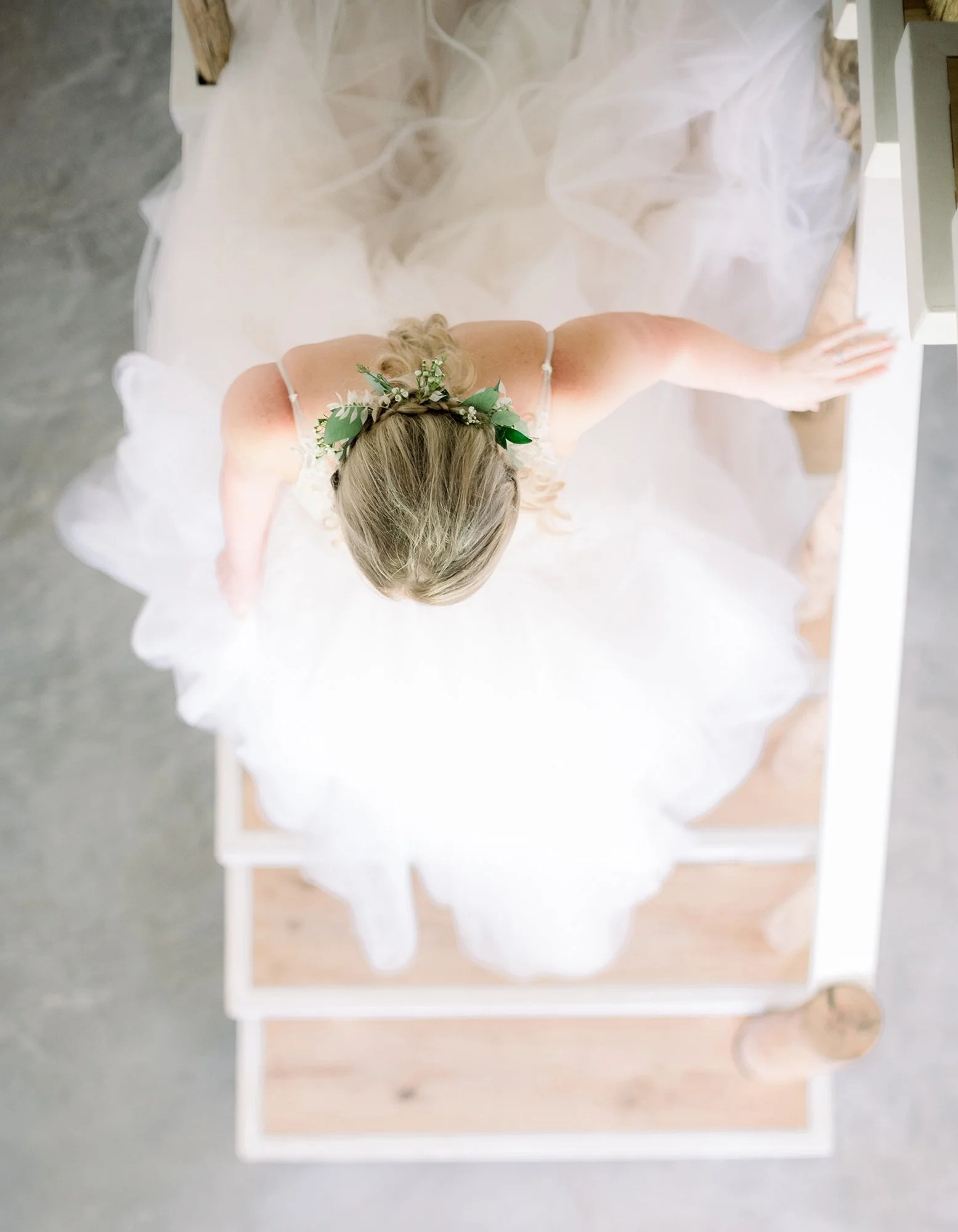 A top-down view of a bride in a wedding dress with a floral headpiece, sitting on a wooden bench, with her arm resting on a white surface.