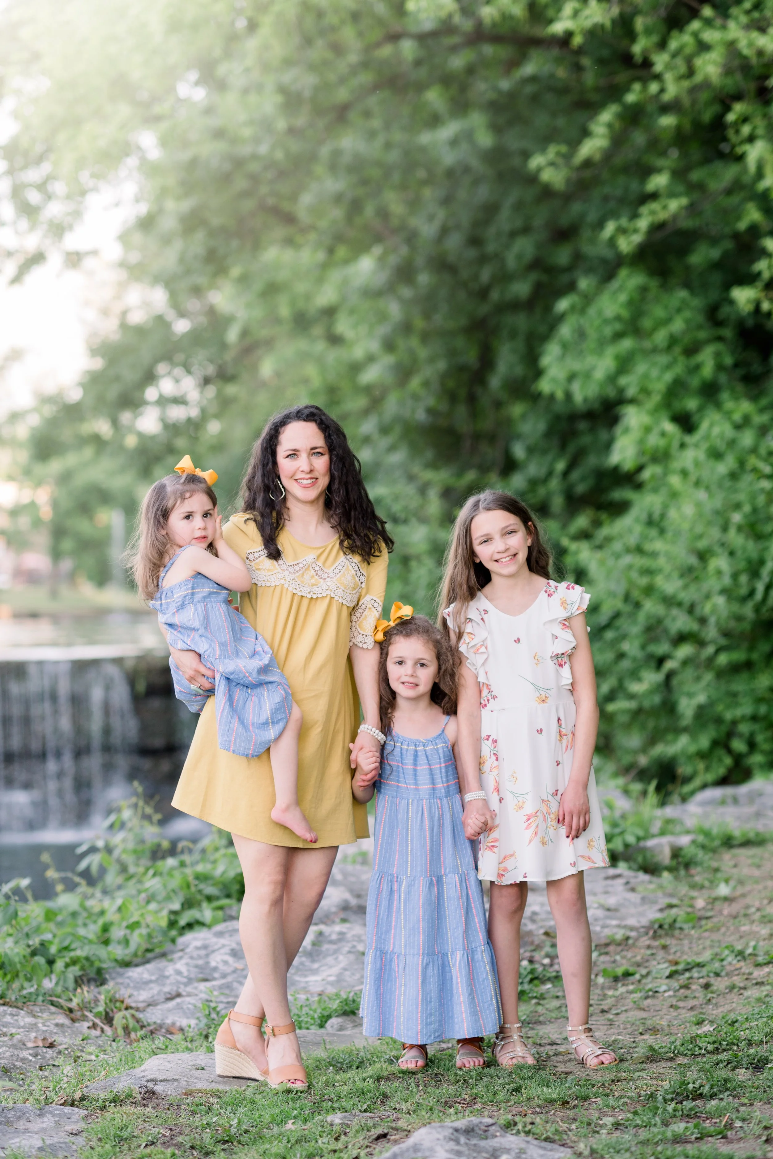 A woman and three girls standing outdoors near a waterfall and lush green trees, holding hands and smiling at the camera.