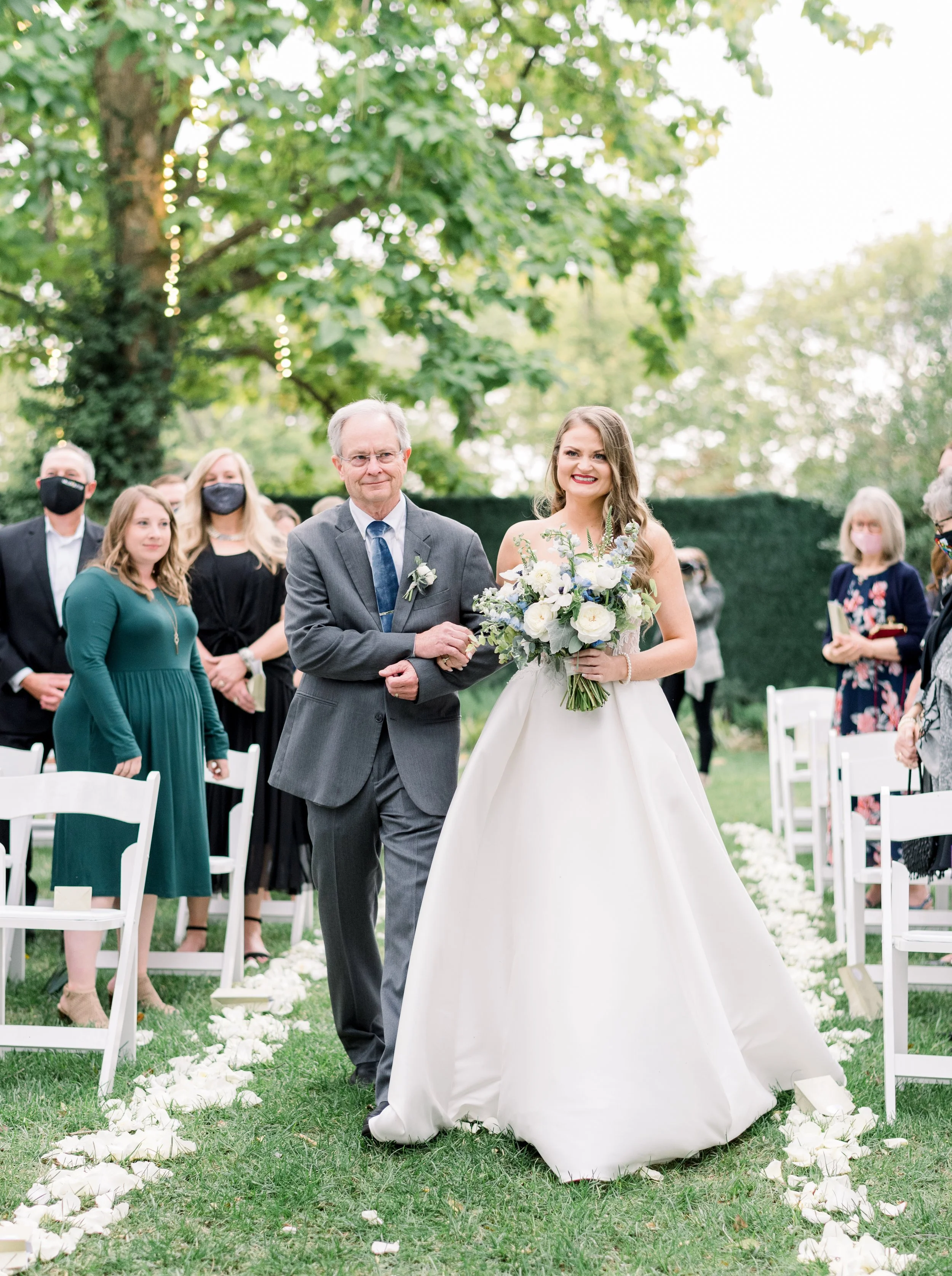 A bride walking down the outdoor wedding aisle with her father, holding a bouquet of white flowers, surrounded by wedding guests seated on white chairs, on a grassy area with large trees in the background.