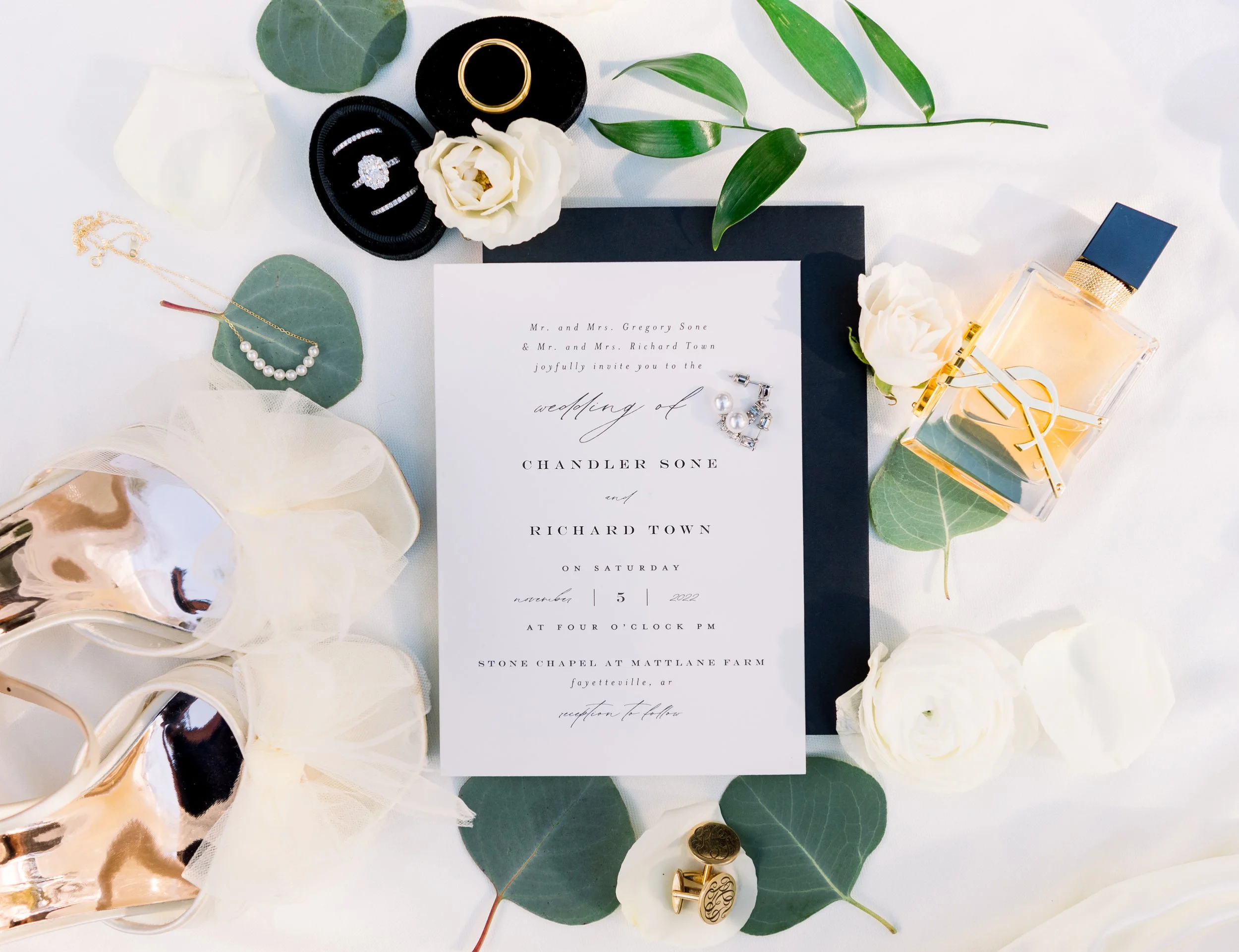 Wedding invitation surrounded by jewelry, flowers, a perfume bottle, and decorative items on a white table.