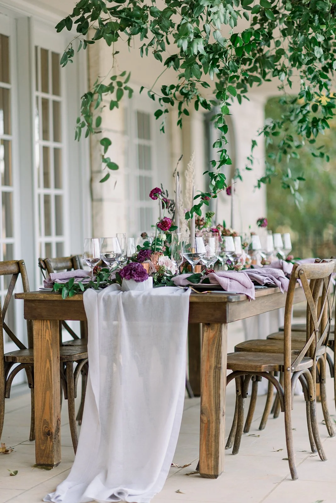 Elegant outdoor dining table decorated with purple and pink flowers, greenery, wine glasses, and lavender napkins, set against a backdrop of a white house with large windows, surrounded by green foliage.