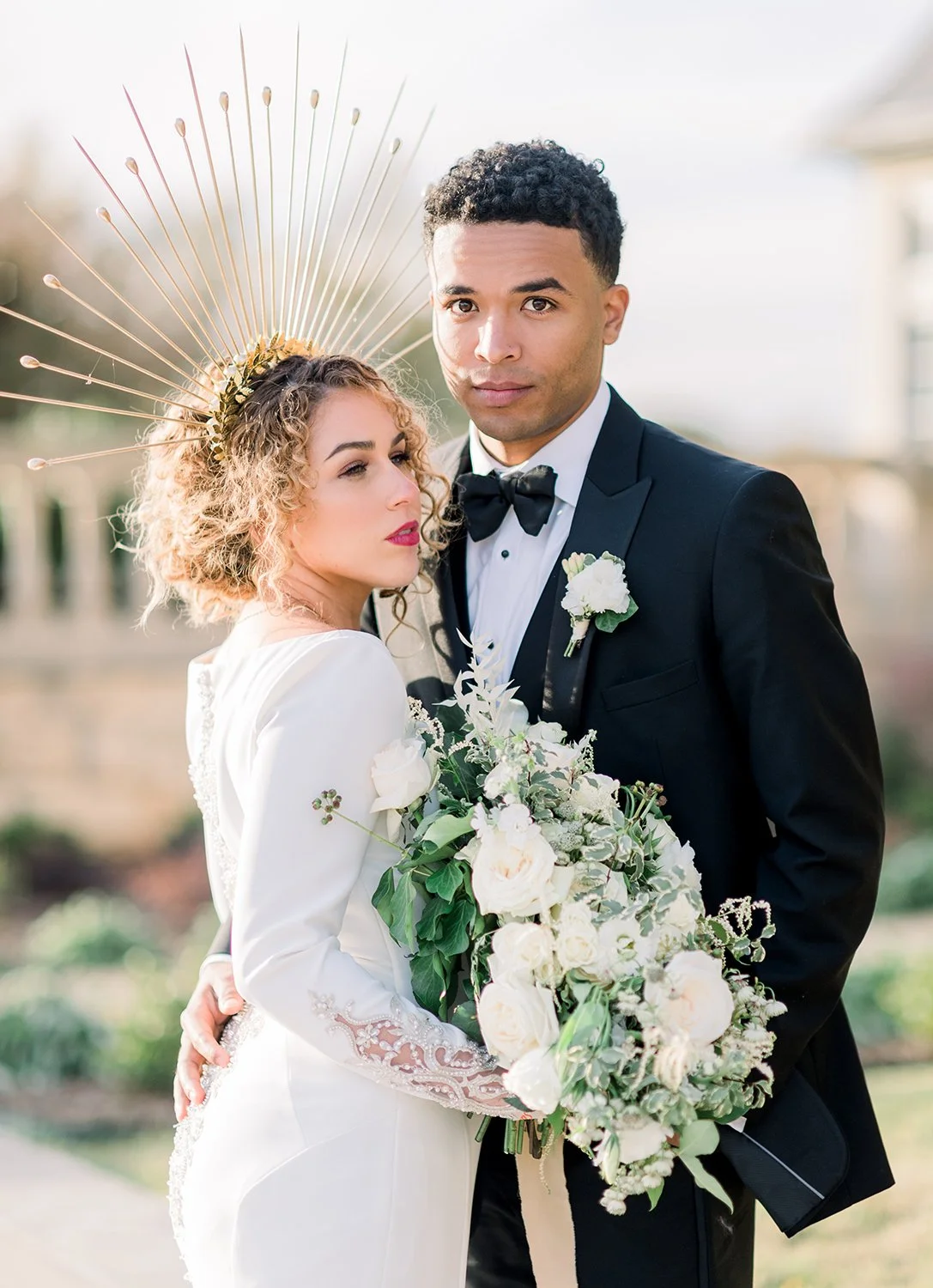 A bride and groom in wedding attire outdoors, with the bride holding a large bouquet of white and green flowers, and the groom wearing a black tuxedo with a bow tie and a white boutonniere. The bride has curly hair and wears a white dress with lace d