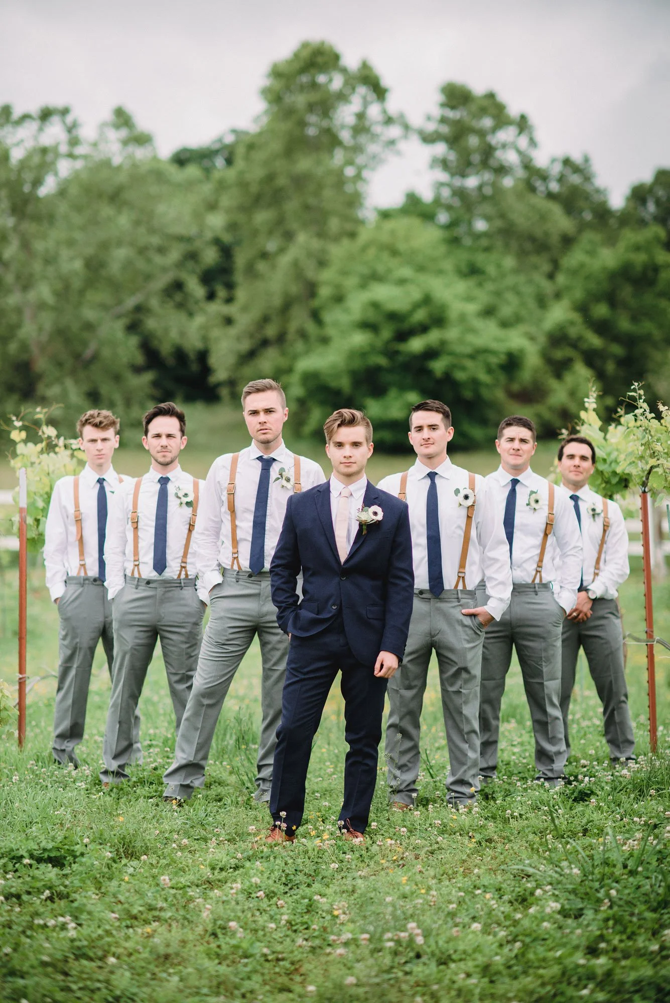 A group of seven men dressed in formal attire, standing outdoors in a green, wooded area. The man in front is wearing a navy suit, while the other six men are wearing white shirts, grey pants, and navy ties with suspenders.