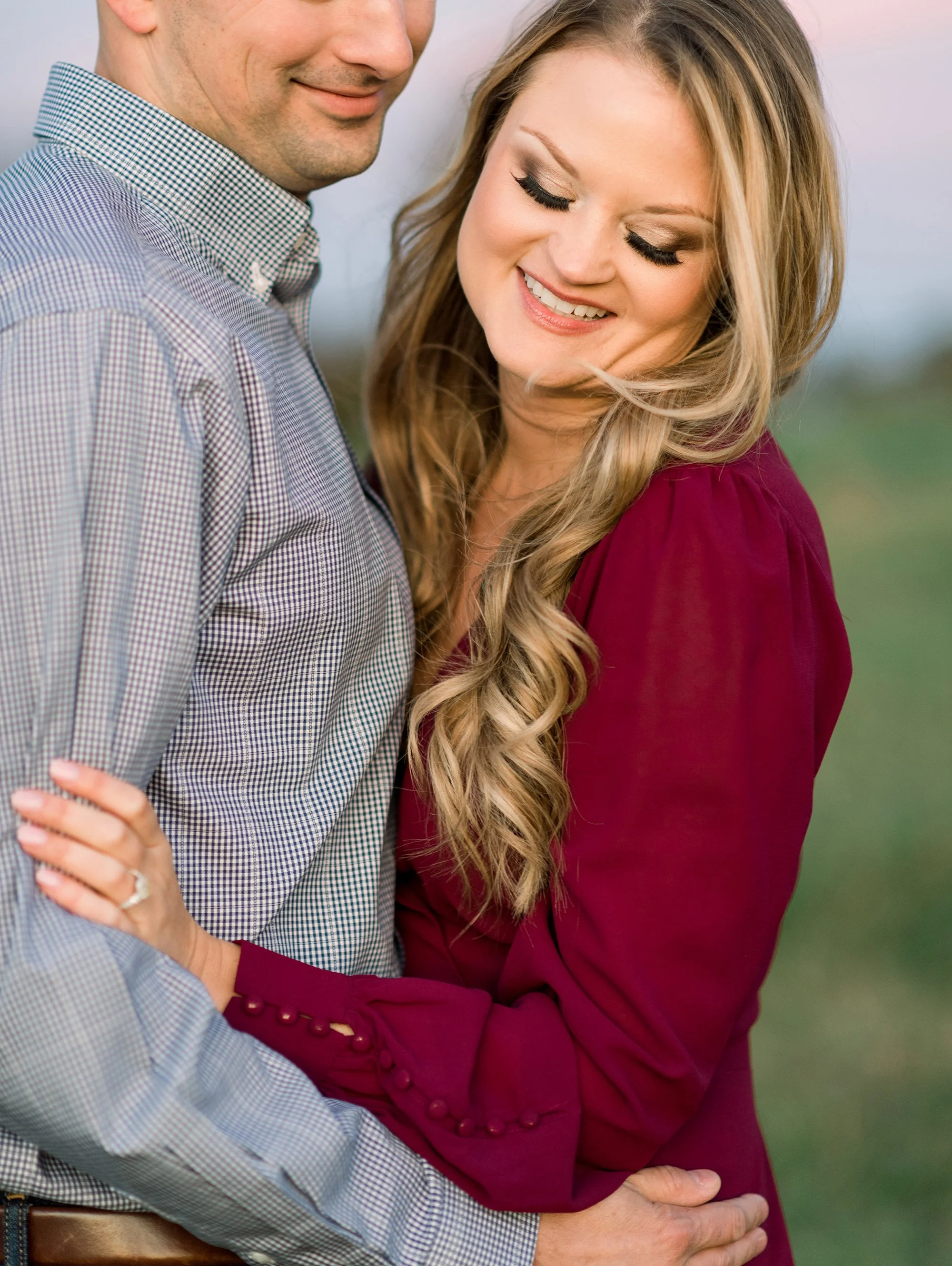 A smiling man and woman embrace outdoors, woman wearing a maroon dress and woman wearing a checkered shirt.