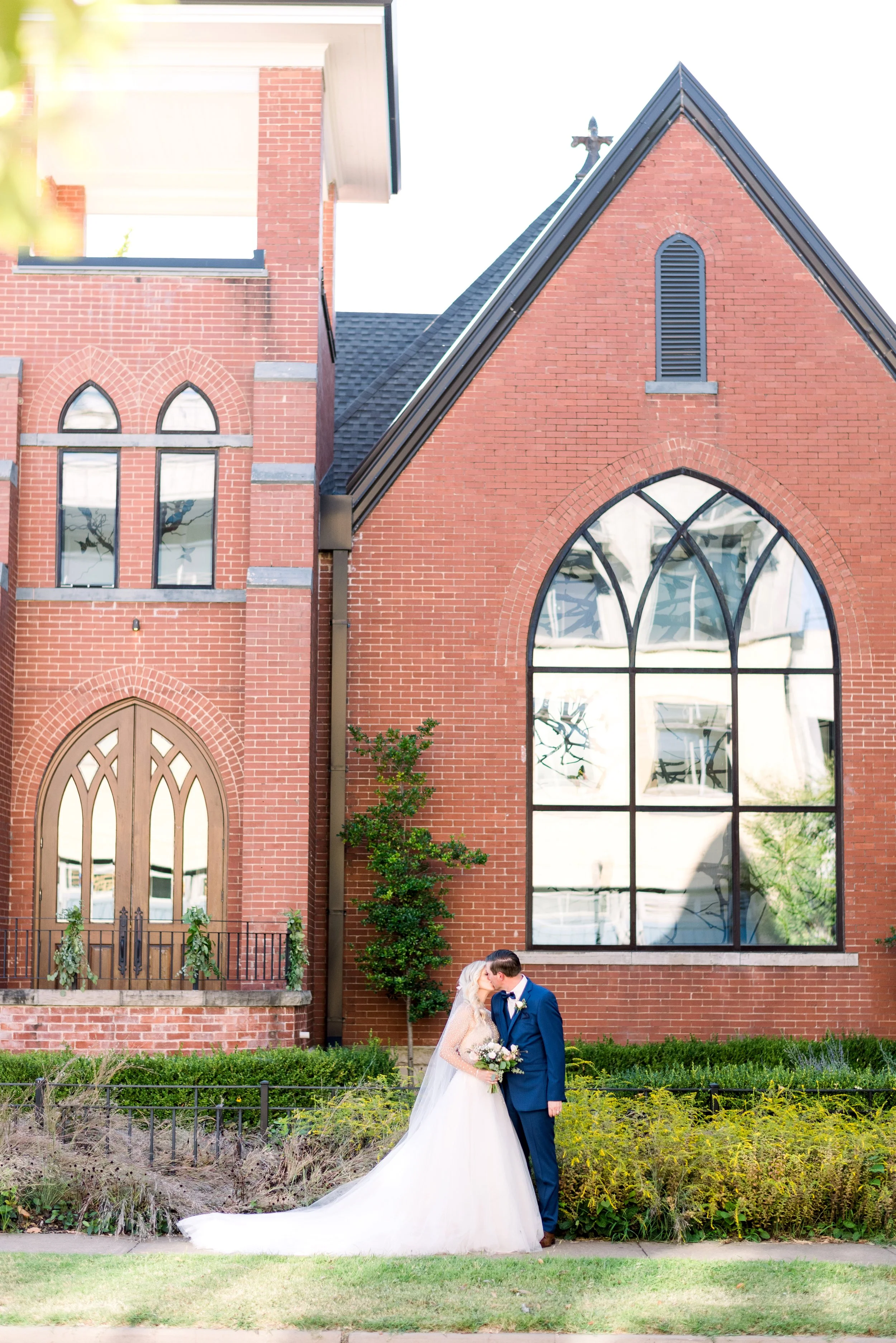 A bride and groom are kissing outdoors in front of a large red brick church with tall stained-glass windows and an arched wooden door, during their wedding.