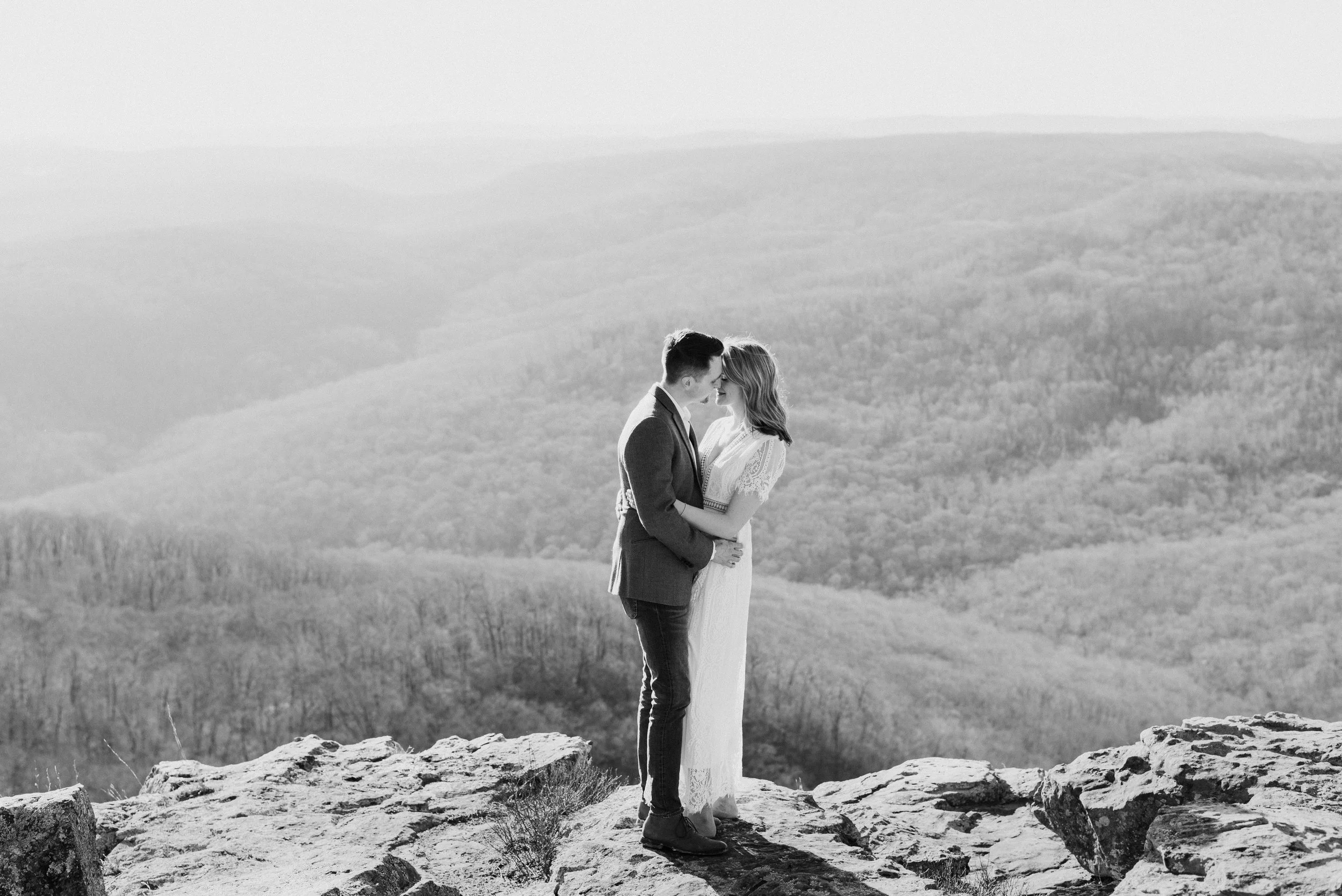 A black-and-white photo of a couple standing on rocky terrain, embracing, with a vast, hilly landscape in the background.