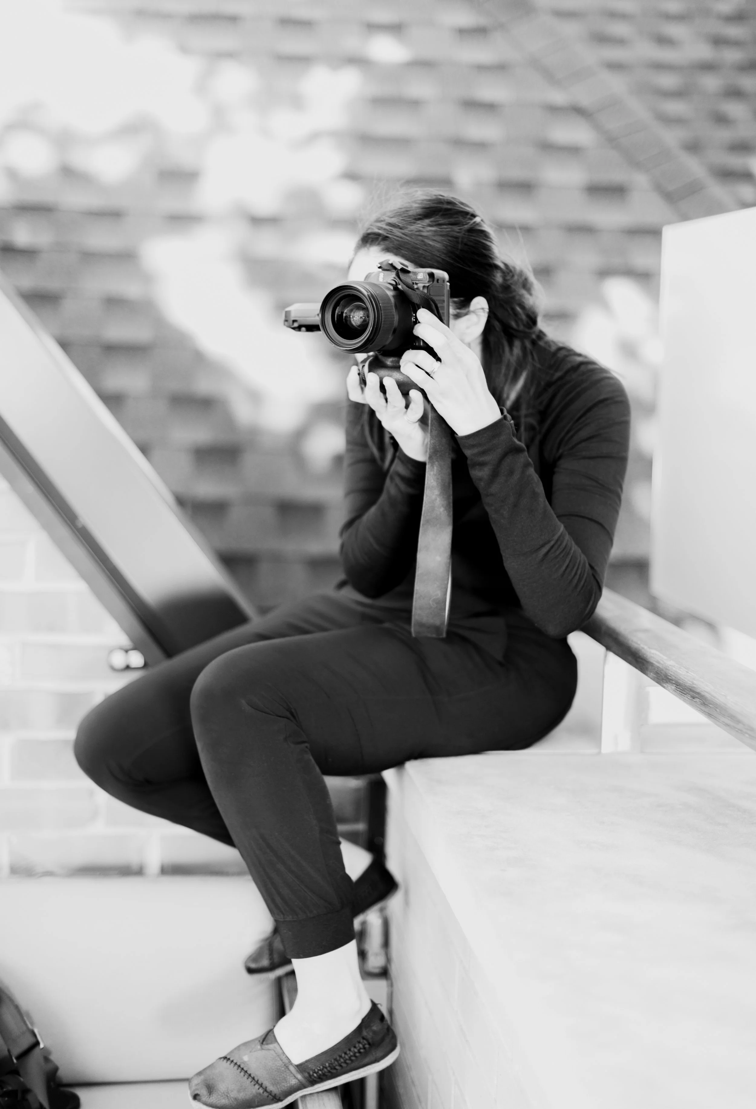 A woman sitting outdoors on steps, holding a camera up to her face, taking a photo. She is dressed in dark clothing and sneakers, with a camera strap around her neck.