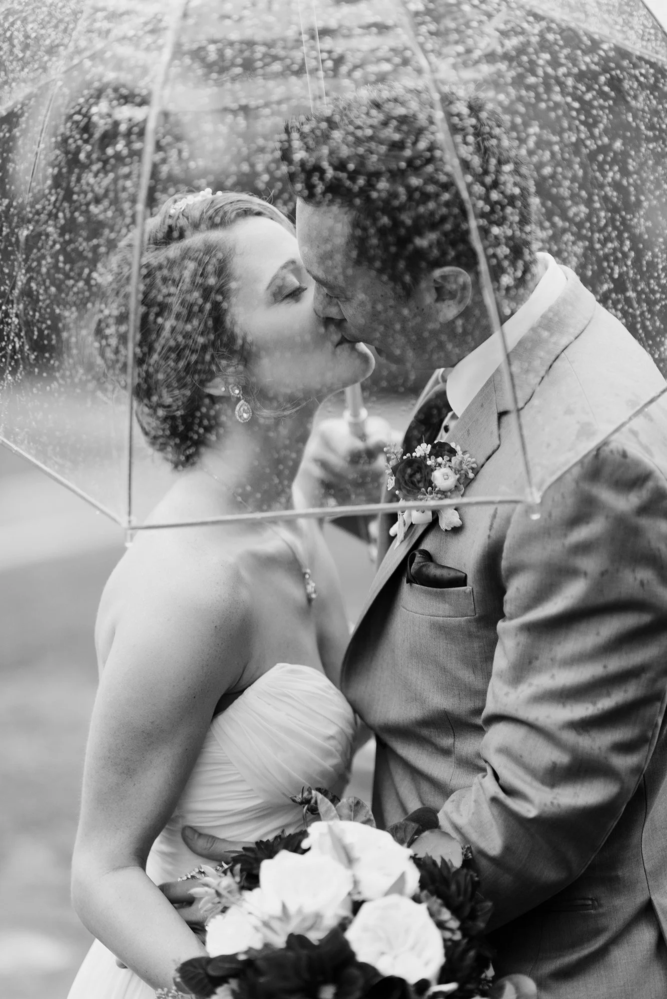 A black-and-white photo of a bride and groom kissing under an umbrella on their wedding day, with raindrops visible on the umbrella.
