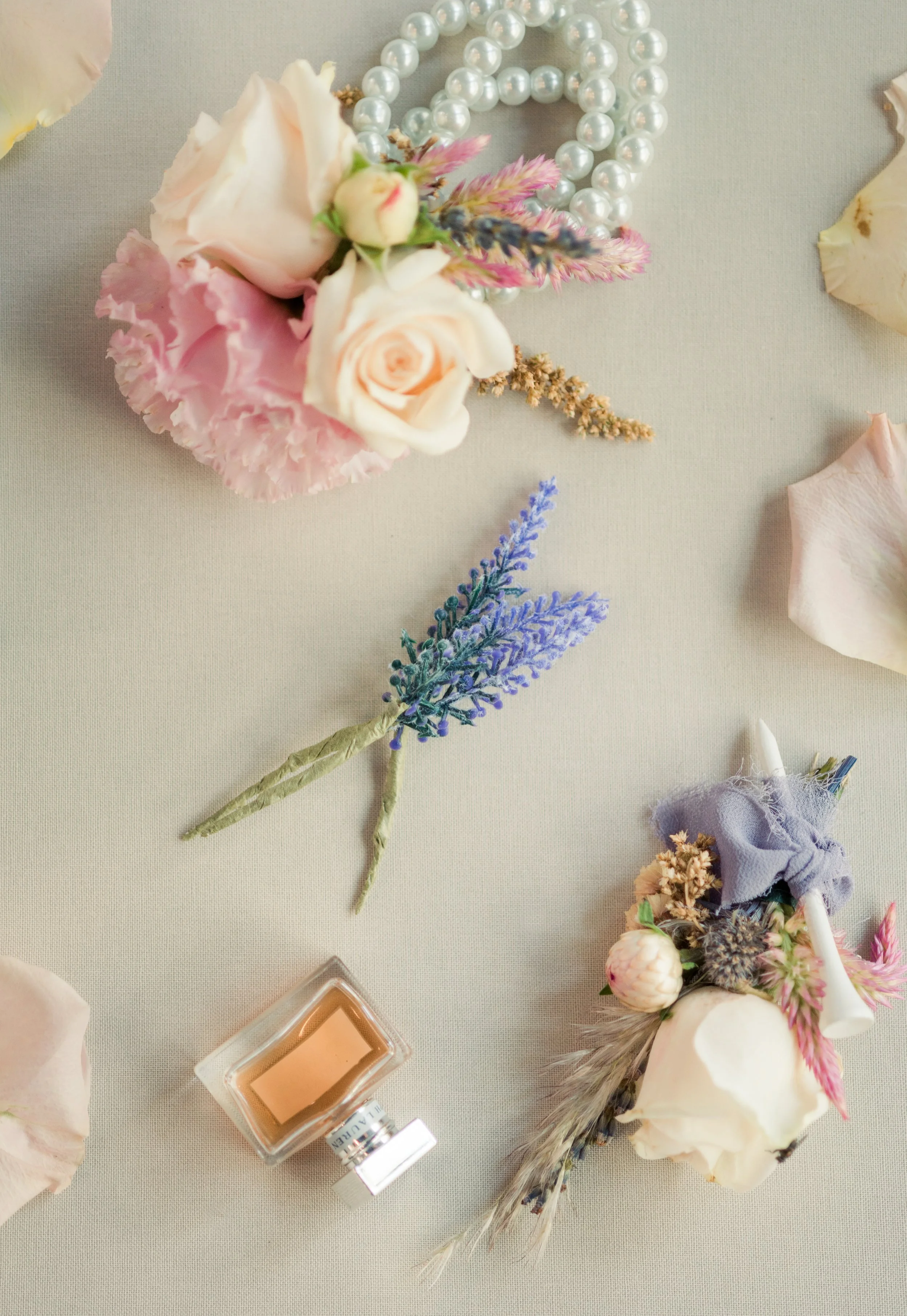 Flat lay of floral boutonnieres, pearl necklace, perfume bottle, and scattered rose petals on a neutral surface.