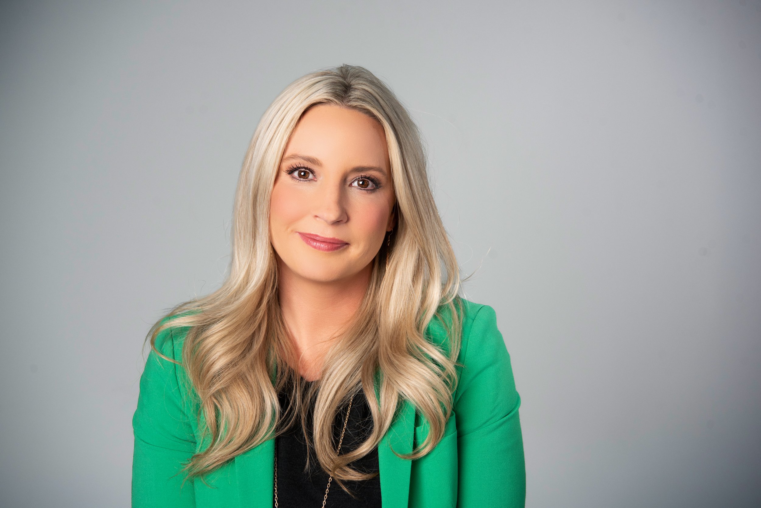 A woman with long blonde hair wearing a green blazer and a black top, smiling softly against a light gray background.