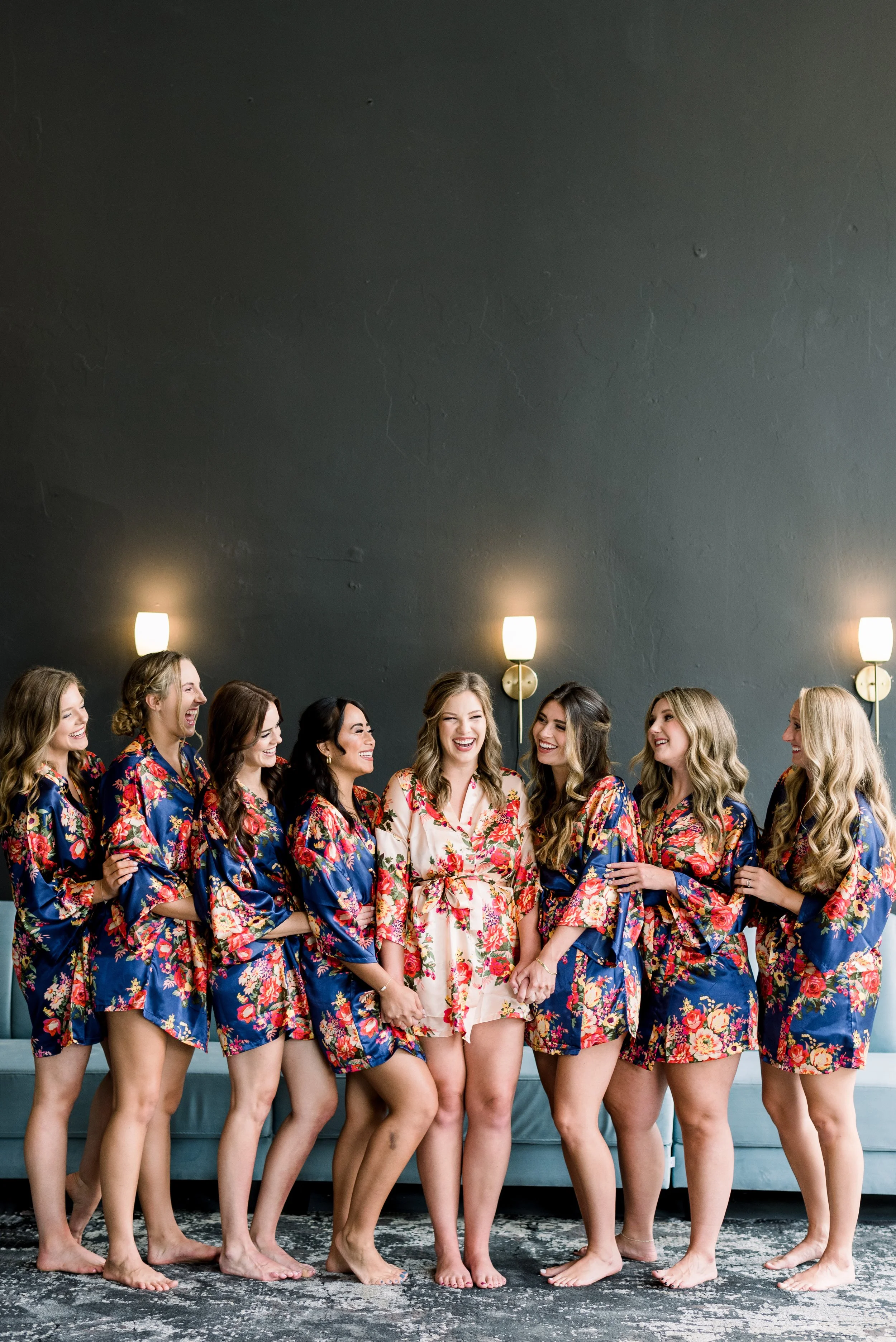 Group of women in floral robes smiling and laughing together in a room with dark wall and wall-mounted lamps.